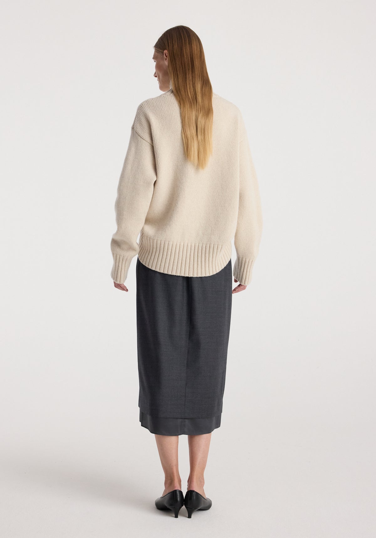 Rear view of a woman wearing a cream textured sweater paired with a dark grey melange Boxer detailed skirt | dark grey melange, standing against a plain white background.