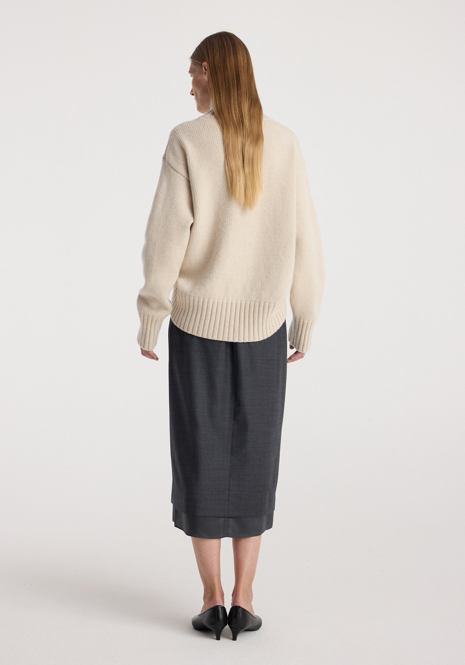 Rear view of a woman wearing a cream textured sweater paired with a dark grey melange Boxer detailed skirt | dark grey melange, standing against a plain white background.