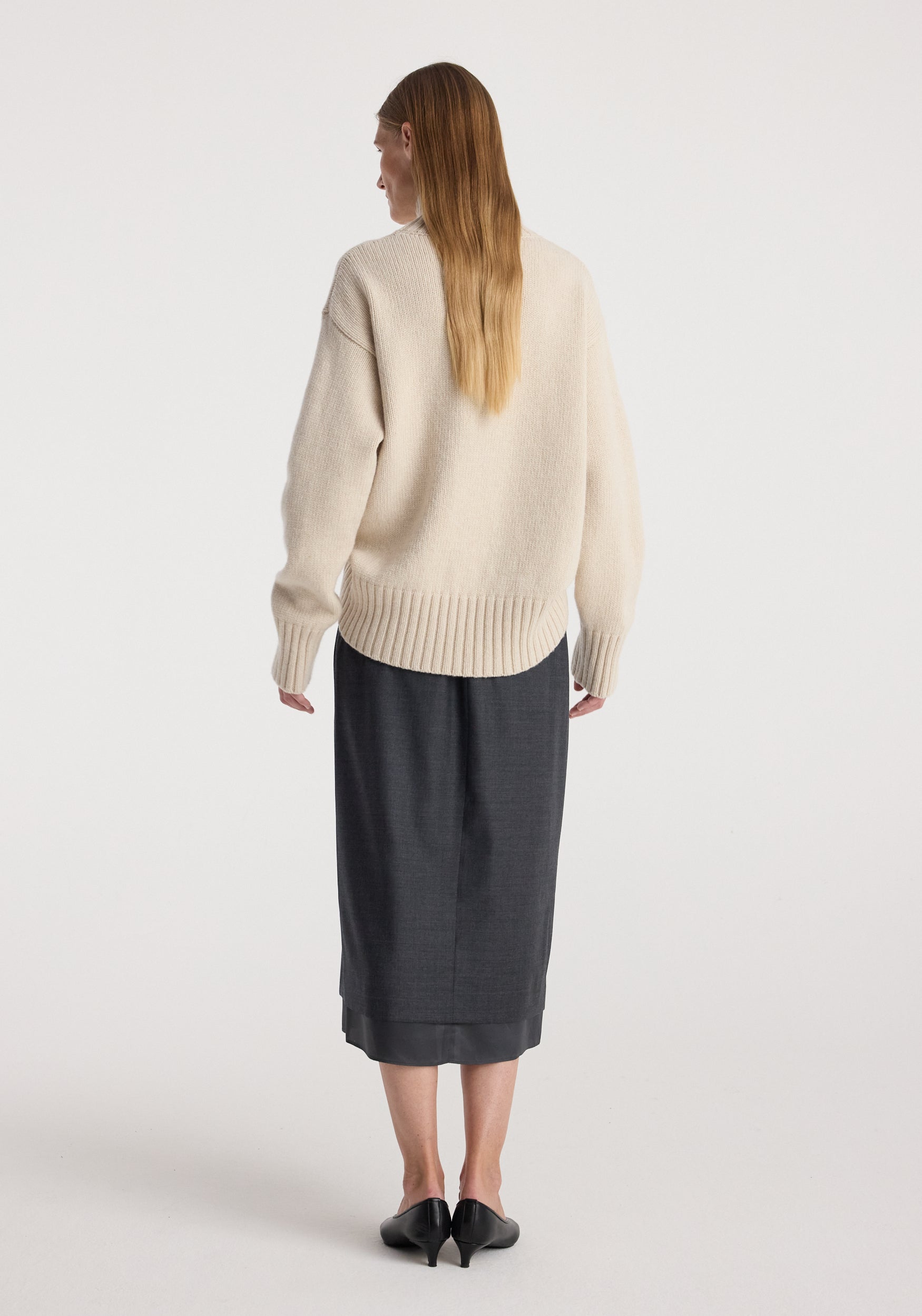 Rear view of a woman wearing a cream textured sweater paired with a dark grey melange Boxer detailed skirt | dark grey melange, standing against a plain white background.