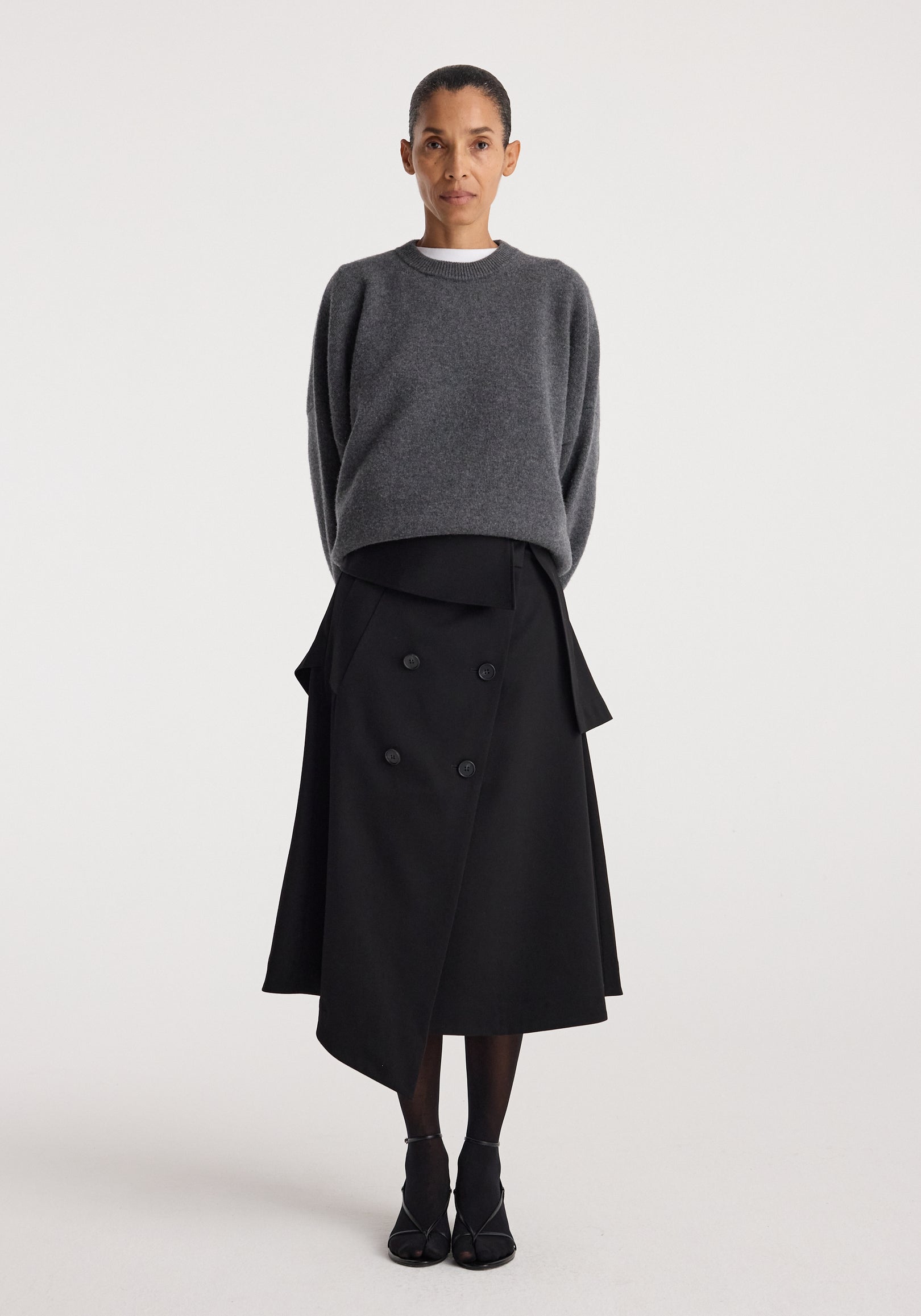 A woman wearing a black Deconstructed trench skirt | black with an asymmetrical double-breasted design paired with a dark gray oversized sweater and black shoes, standing against a plain white background.