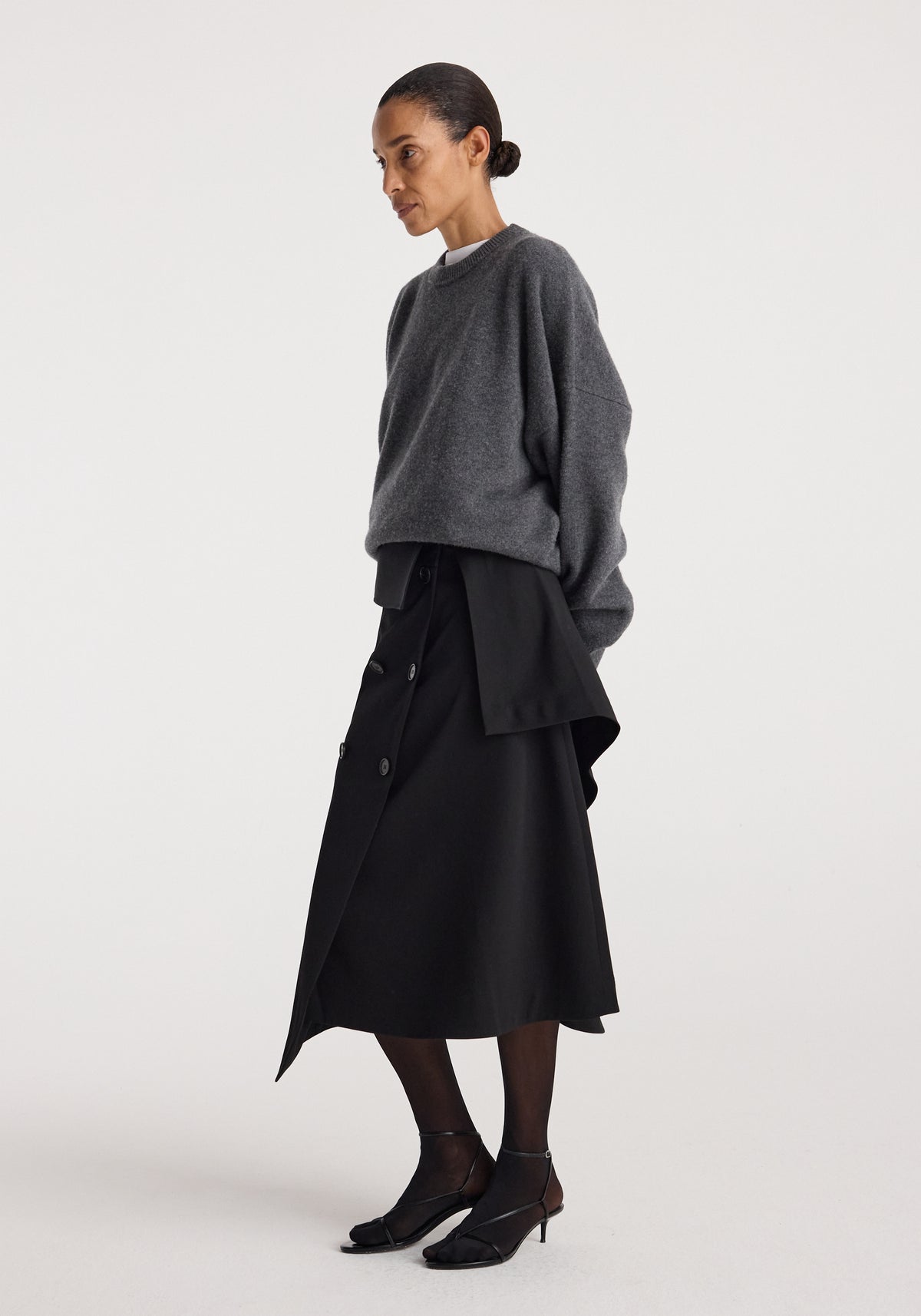 Woman wearing a loose dark grey sweater paired with a black asymmetrical buttoned skirt featuring layered fabric design and black sheer tights, styled with black kitten heels, showcasing the Deconstructed trench skirt | black.