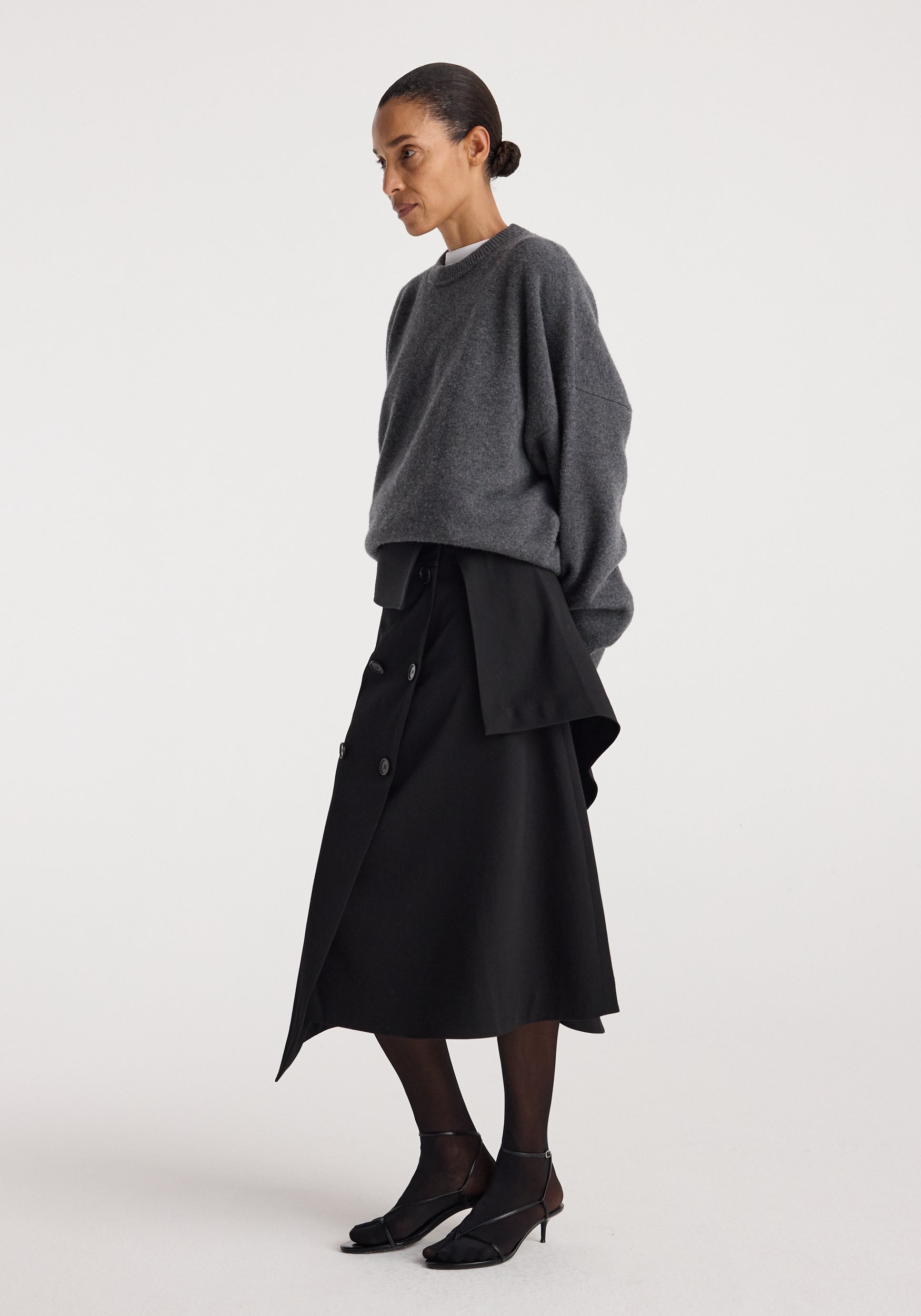 Woman wearing a loose dark grey sweater paired with a black asymmetrical buttoned skirt featuring layered fabric design and black sheer tights, styled with black kitten heels, showcasing the Deconstructed trench skirt | black.