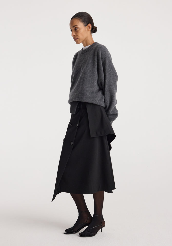 A woman wearing a black Deconstructed trench skirt | black with an asymmetrical double-breasted design paired with a dark gray oversized sweater and black shoes, standing against a plain white background.