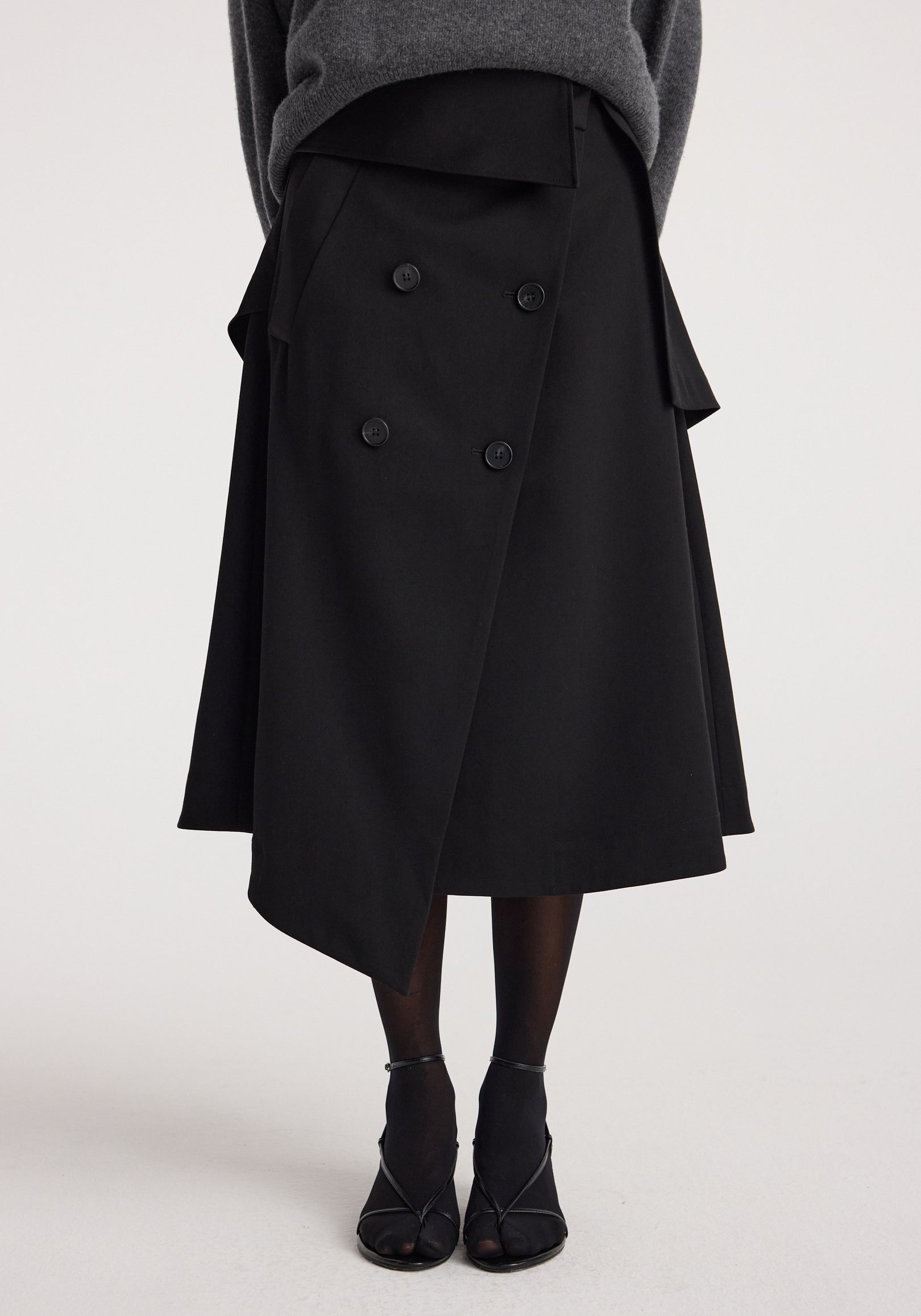 Woman wearing a black Deconstructed trench skirt | black with asymmetrical hem and double-breasted button detail paired with black tights and open-toe heels.