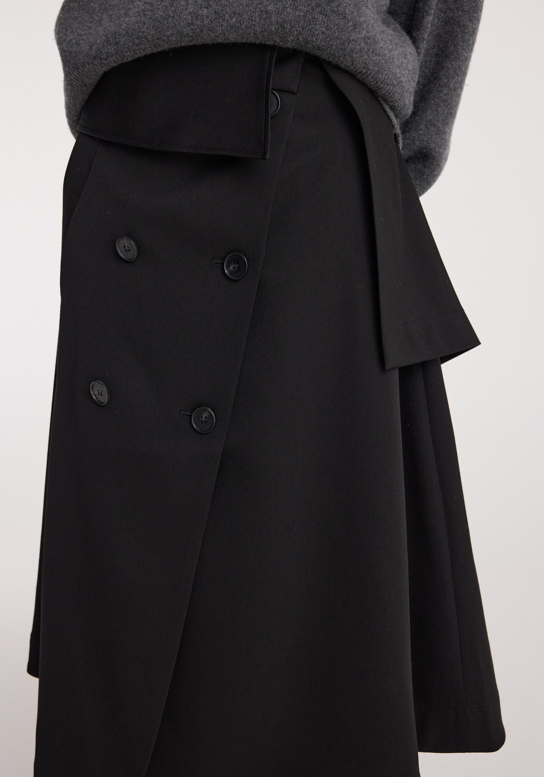 Close-up of a black Deconstructed trench skirt | black featuring an asymmetrical wrap design with button details, worn with a gray sweater.