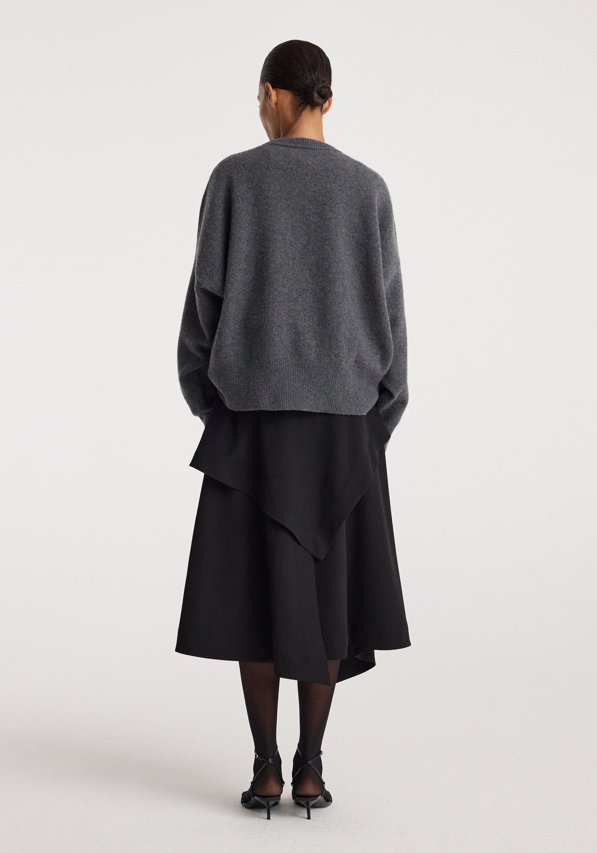 Woman wearing a loose grey sweater paired with a layered black skirt viewed from the back, showcasing the Deconstructed trench skirt | black with an asymmetrical hemline.