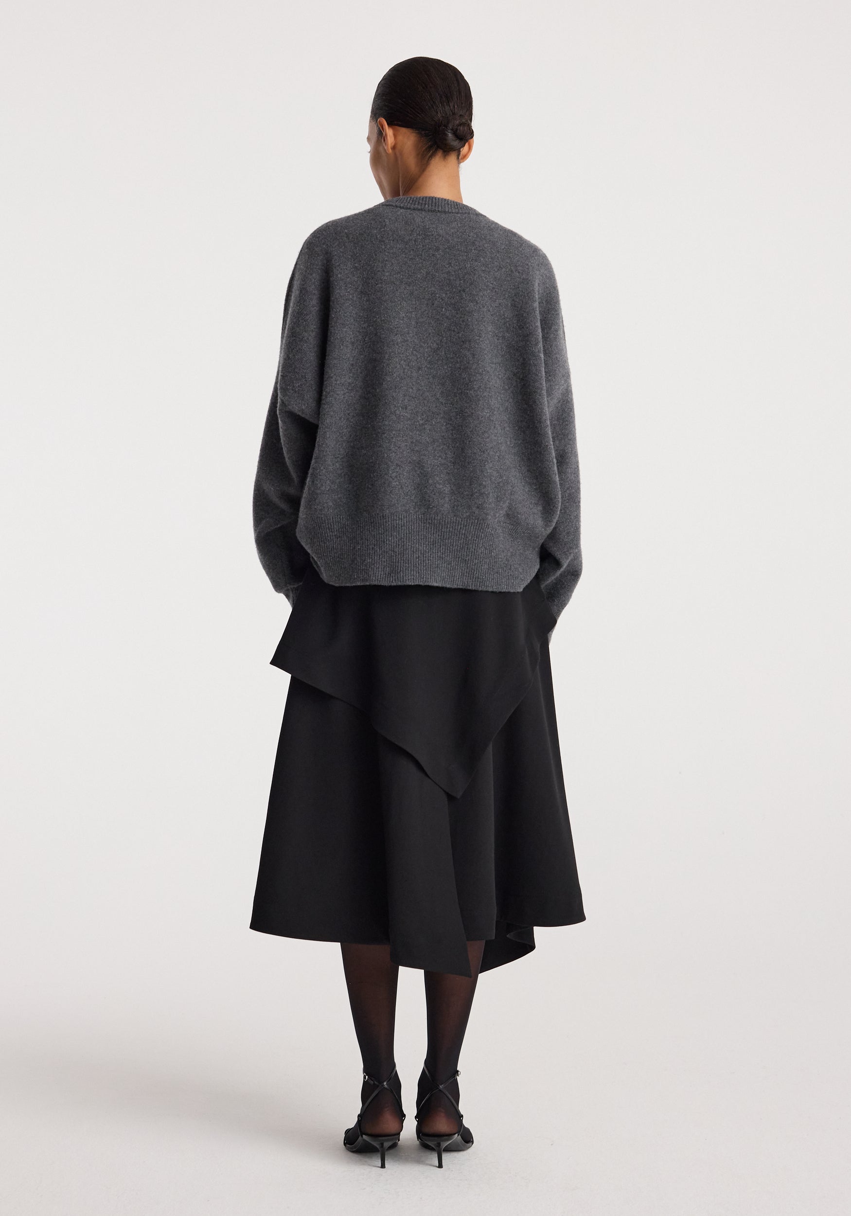 Woman wearing a loose grey sweater paired with a layered black skirt viewed from the back, showcasing the Deconstructed trench skirt | black with an asymmetrical hemline.