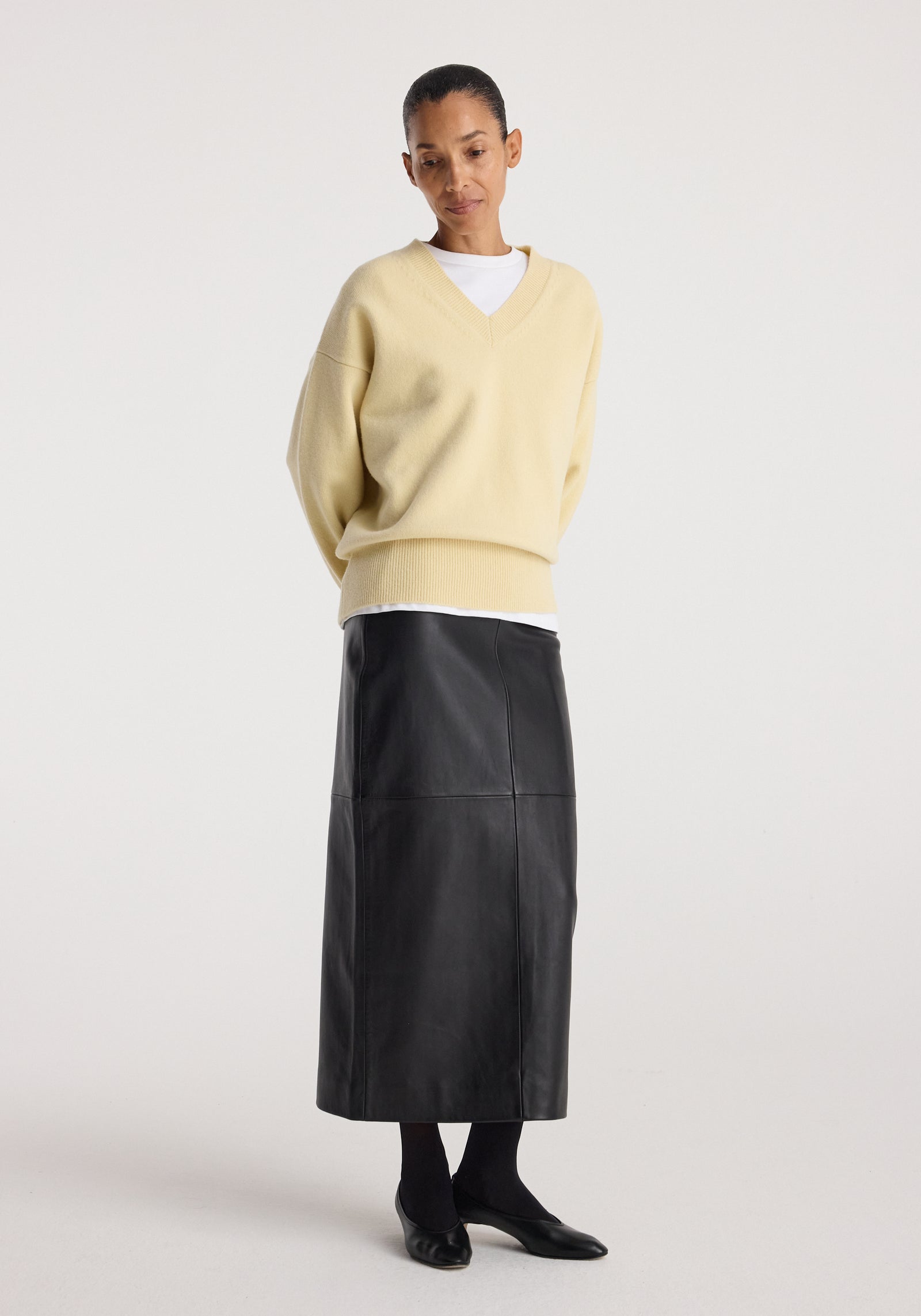 Woman wearing a long black skirt paired with a beige sweater and black shoes standing against a plain background featuring the elongated leather skirt | black.