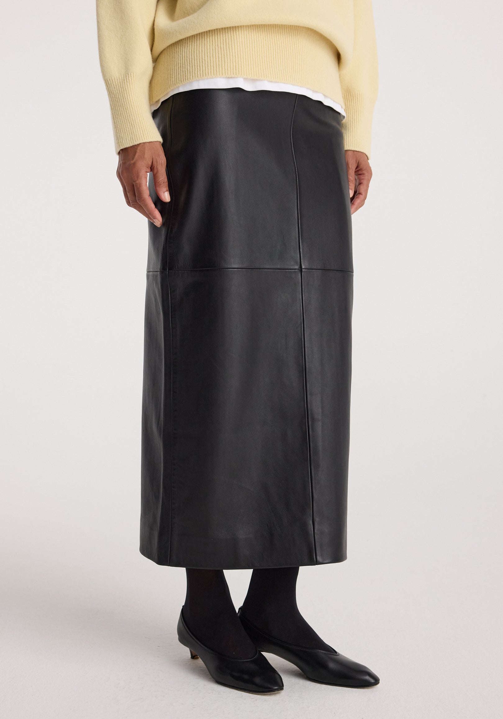 Model wearing an elegant black Elongated leather skirt | black paired with black tights, black low-heeled shoes, and layered tops including a cream sweater over a white shirt