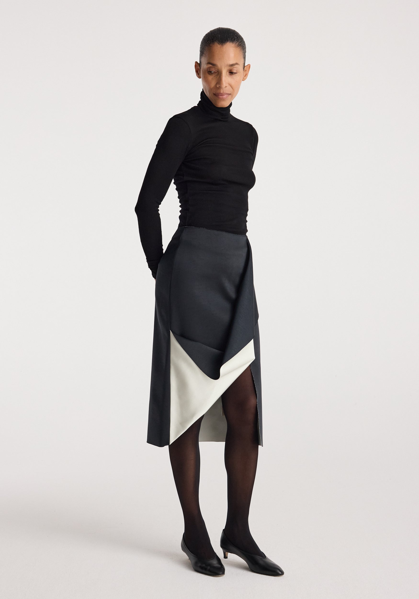 Woman wearing a Sculptural double-satin skirt | black/white with an asymmetrical black and white design, paired with black tights, black heels, and a fitted black turtleneck top.