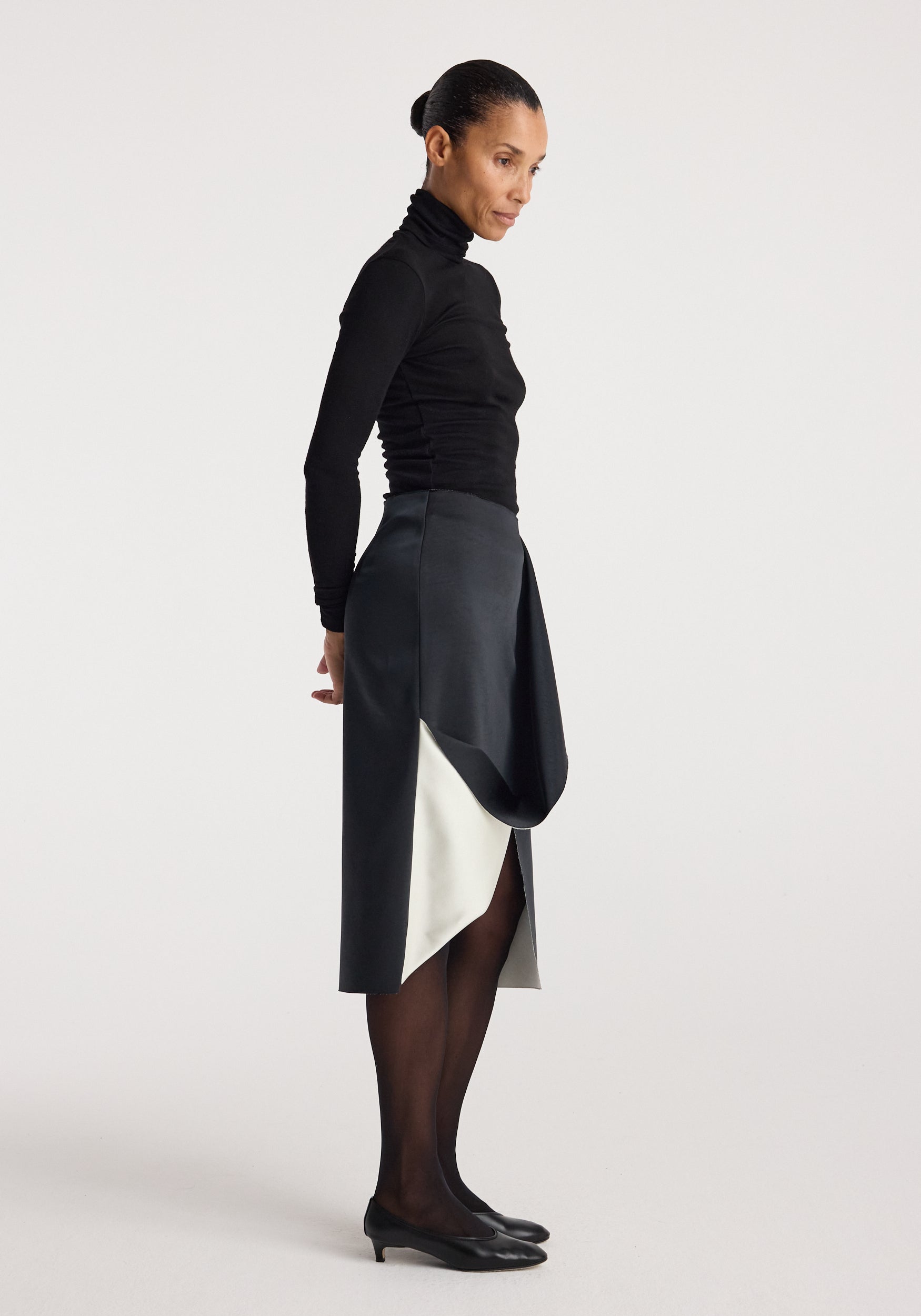 Side profile of a woman wearing a black turtleneck paired with a sculptural double-satin skirt | black/white, featuring layered draped detail and black heels.