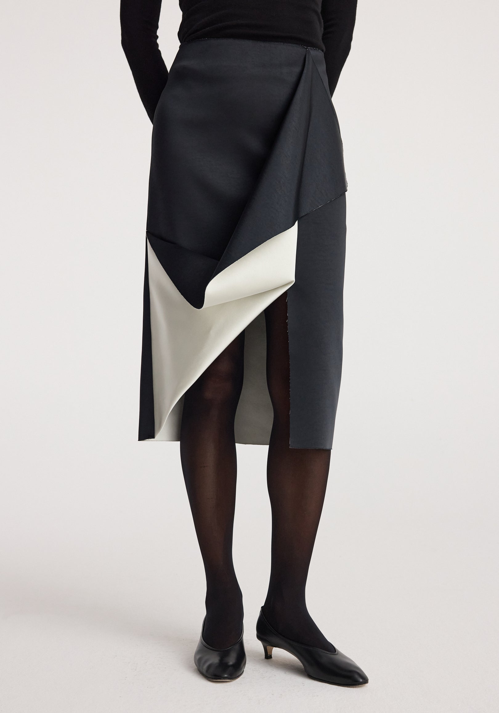 A woman wearing a Sculptural double-satin skirt | black/white featuring an overlapping design with black outer fabric and white inner lining, paired with black tights and black low-heeled shoes.