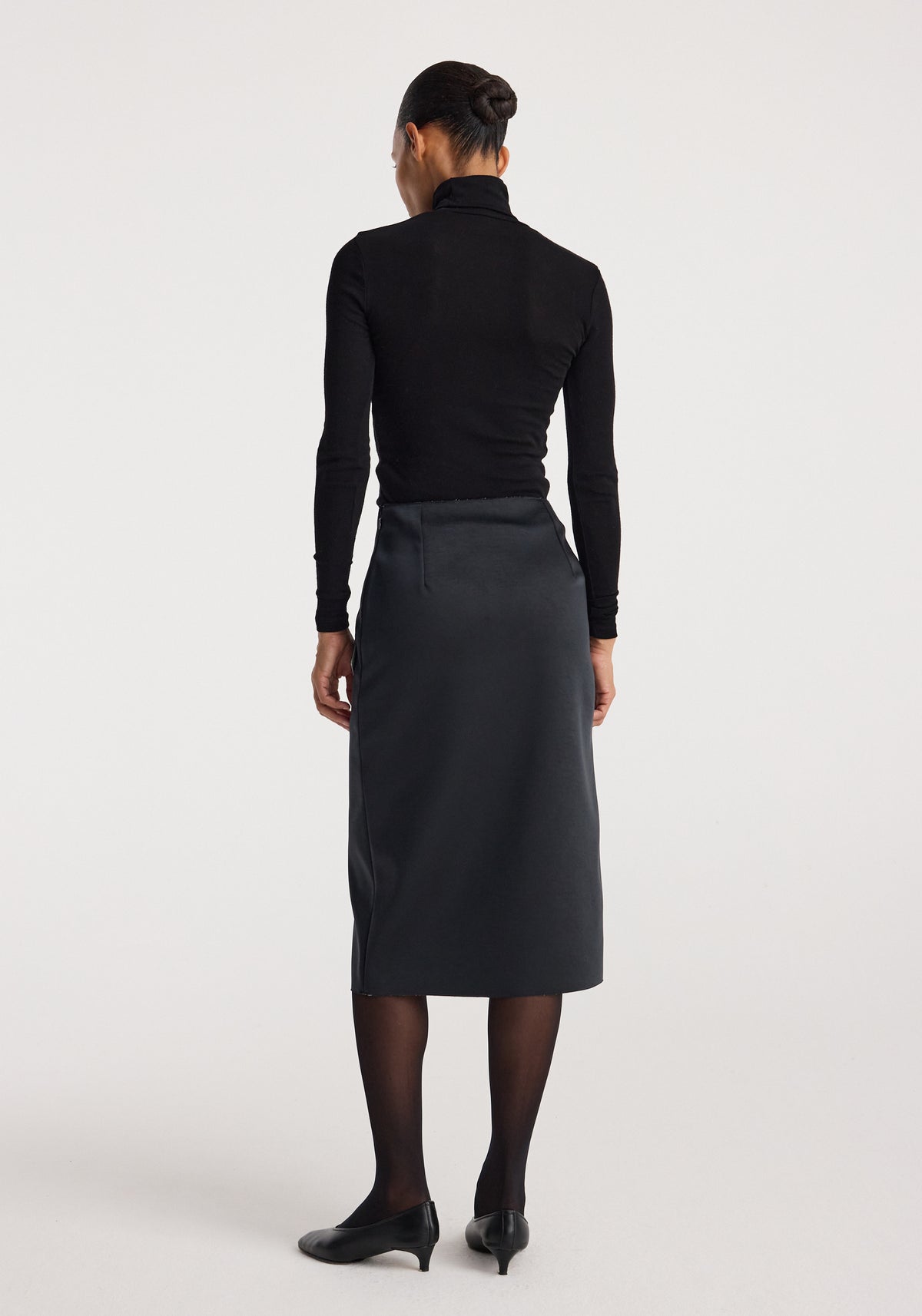 Woman wearing a black long-sleeve top and black heels, viewed from the back, showcasing the sculptural double-satin skirt | black/white with a midi length and sleek fit.
