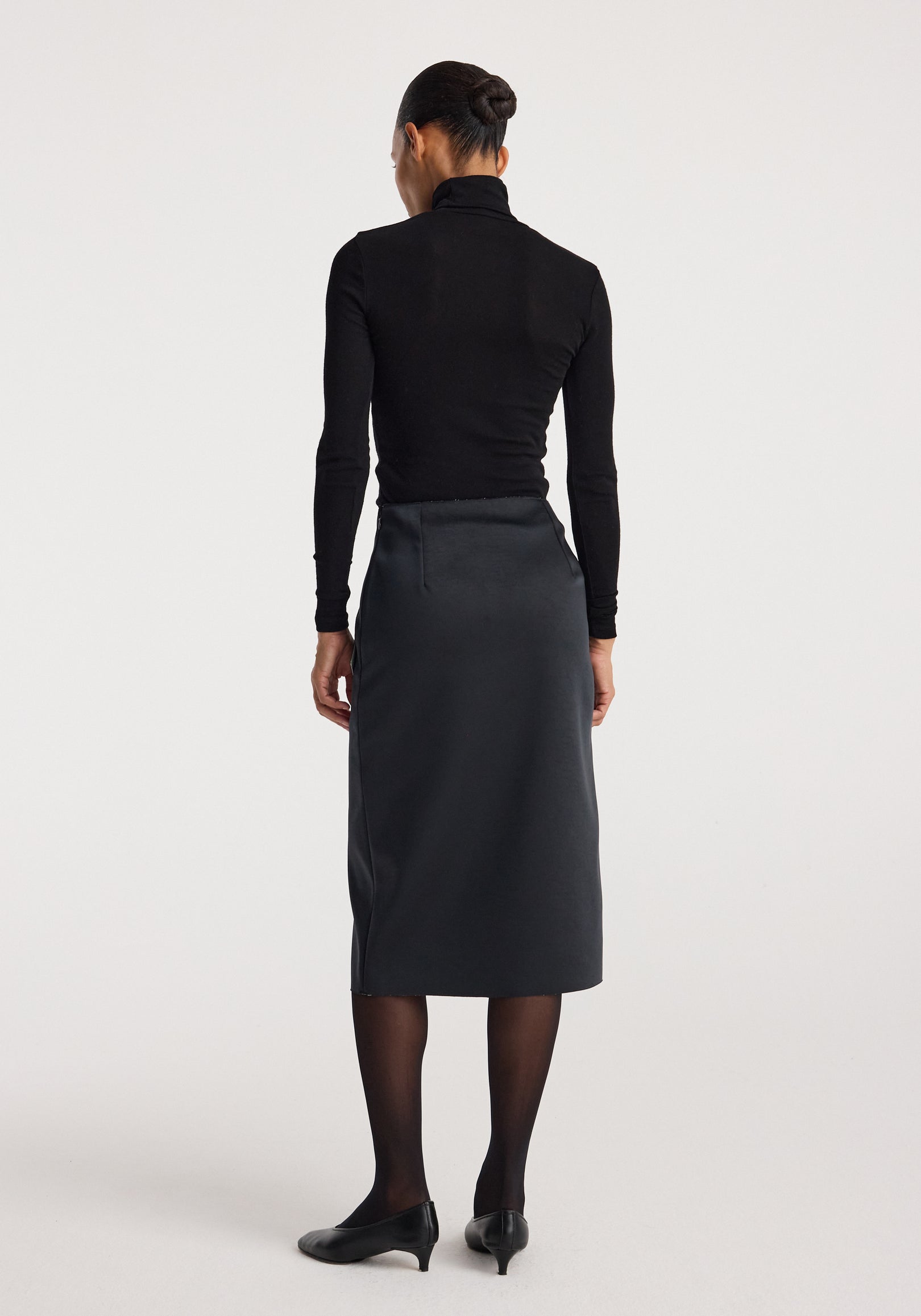 Woman wearing a black long-sleeve top and black heels, viewed from the back, showcasing the sculptural double-satin skirt | black/white with a midi length and sleek fit.