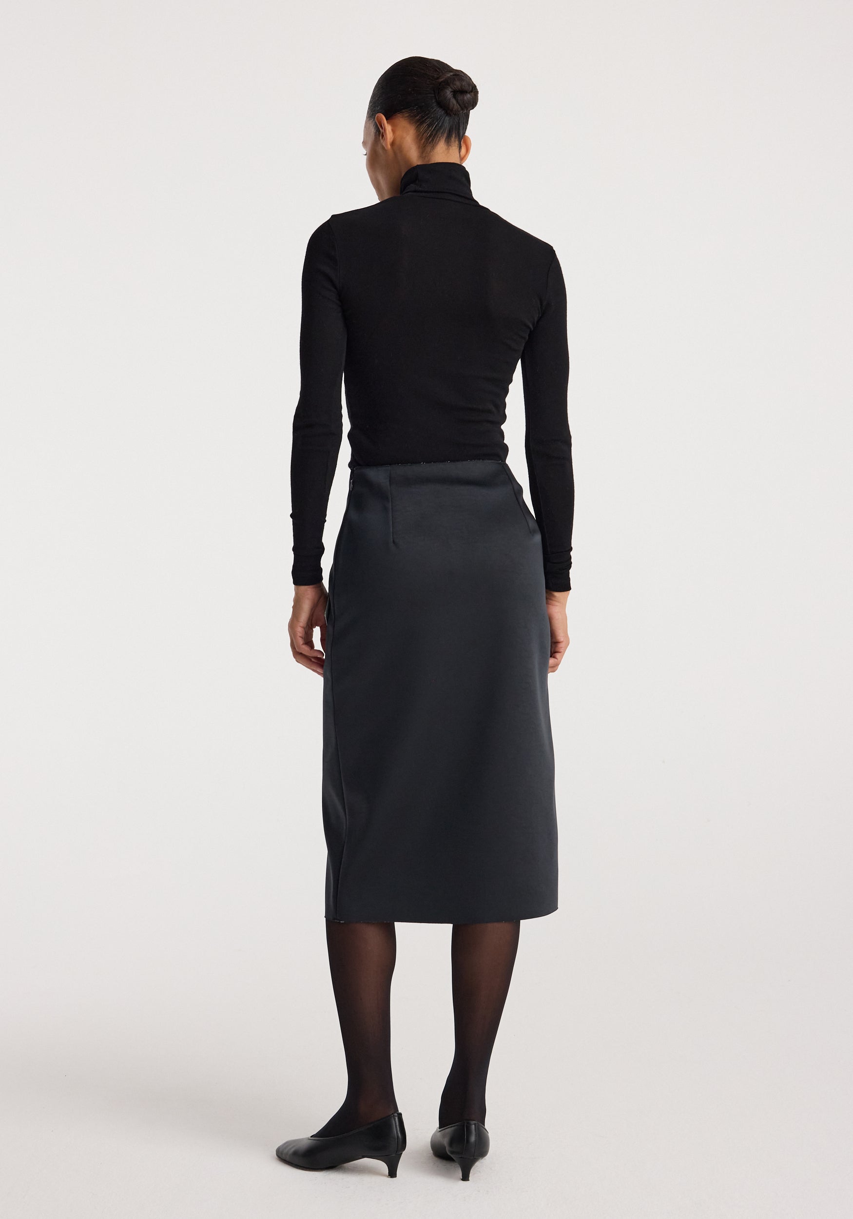 Woman wearing a black long-sleeve top and black heels, viewed from the back, showcasing the sculptural double-satin skirt | black/white with a midi length and sleek fit.