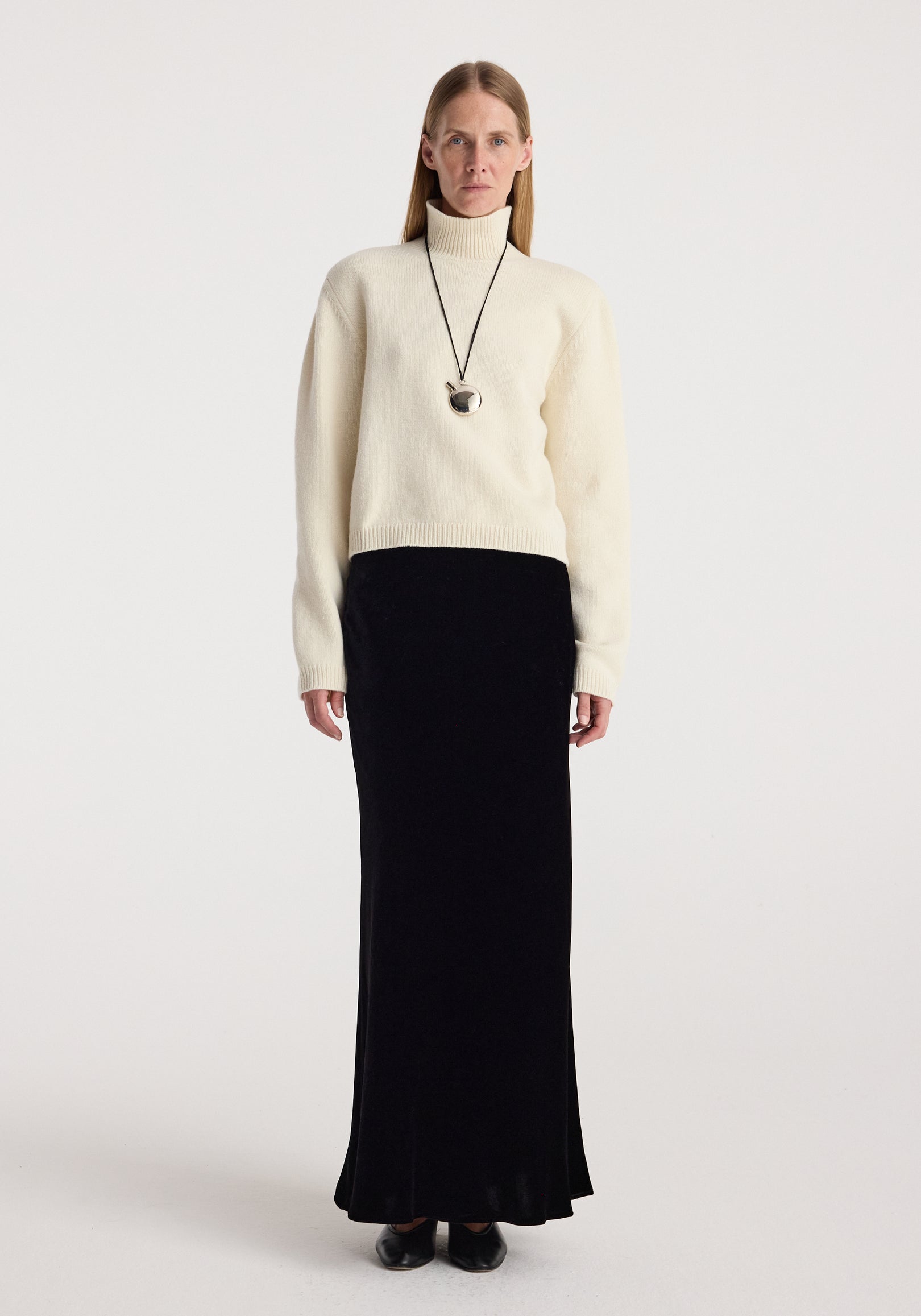 Woman wearing cream turtleneck sweater paired with a long, fitted Fluid velvet skirt | black and black shoes, standing against a plain white background.