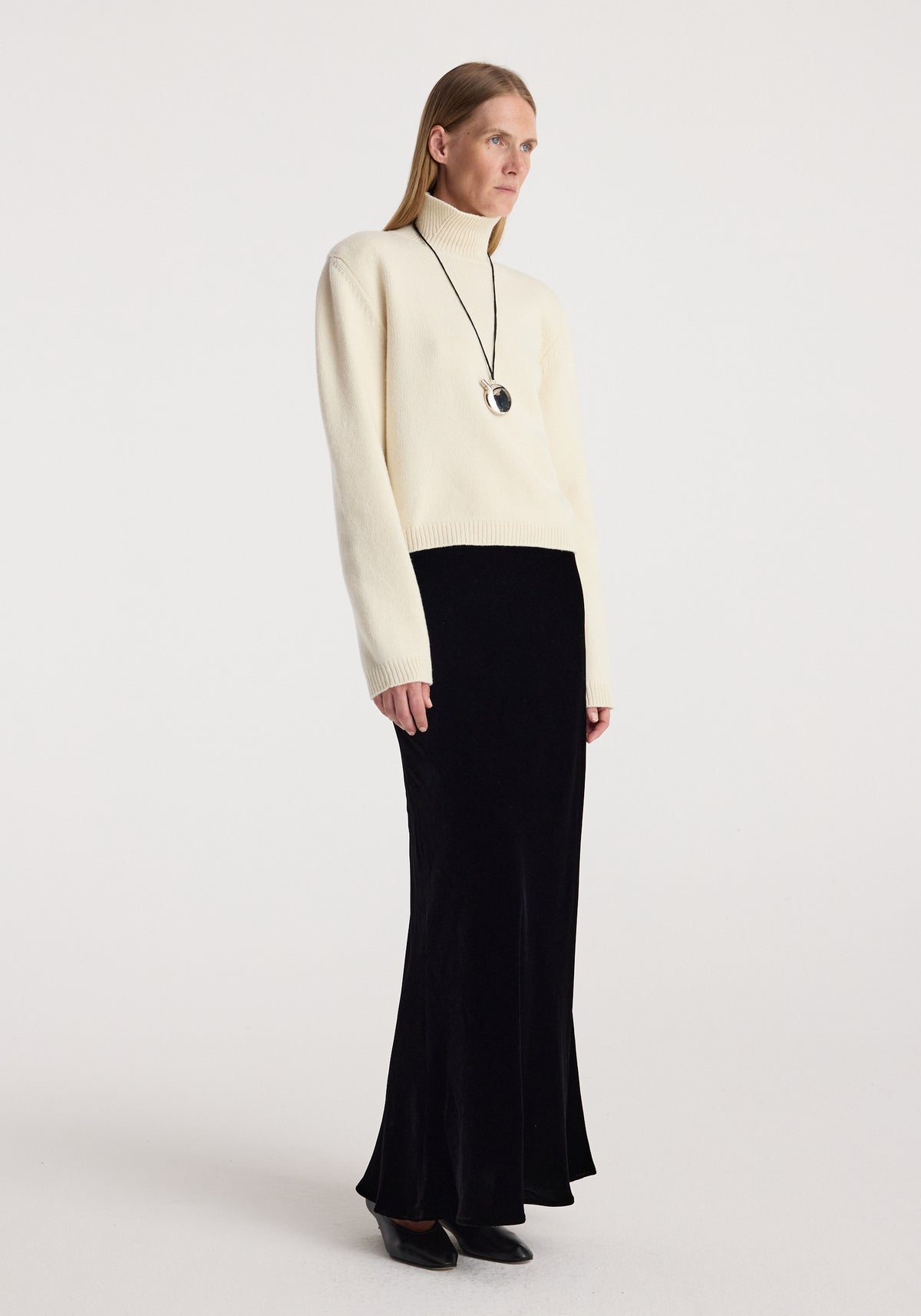 Woman wearing a cream turtleneck sweater paired with a long black velvet skirt, styled with black pointed shoes and a pendant necklace, modeling the Fluid velvet skirt | black.