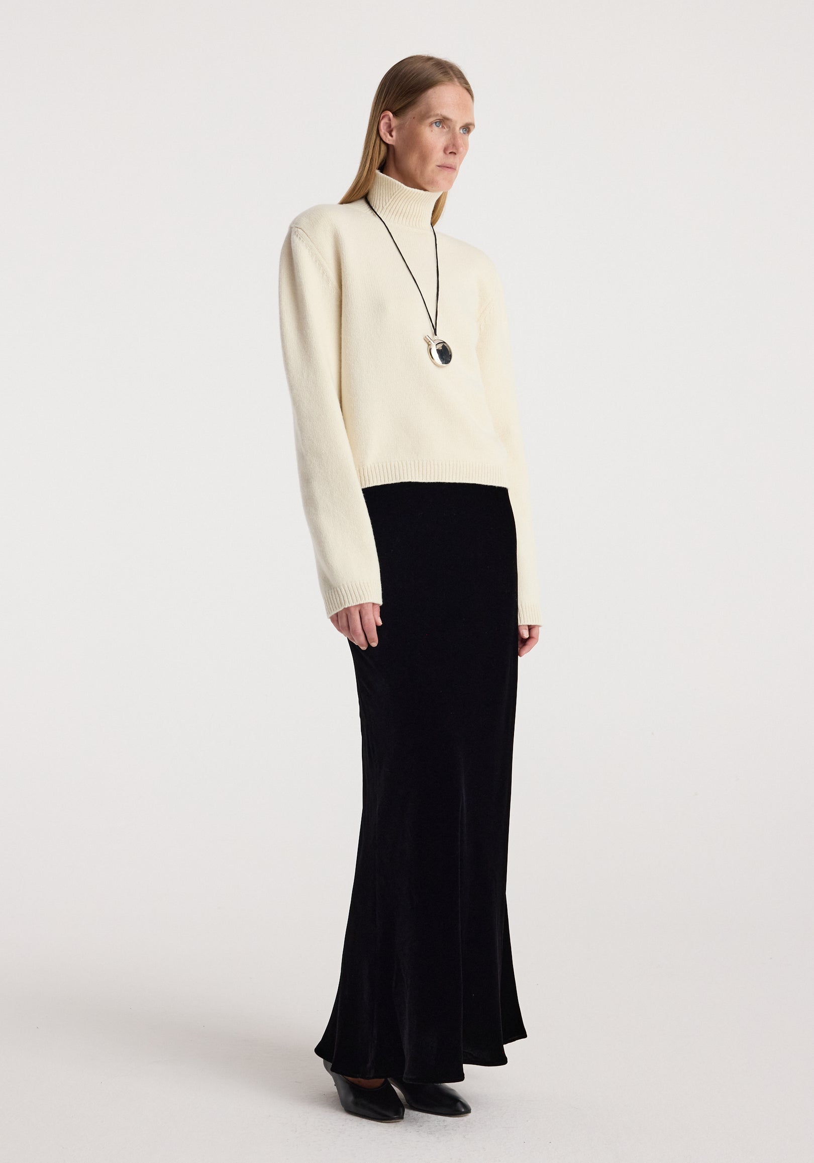 Woman wearing a cream turtleneck sweater paired with a long black velvet skirt, styled with black pointed shoes and a pendant necklace, modeling the Fluid velvet skirt | black.
