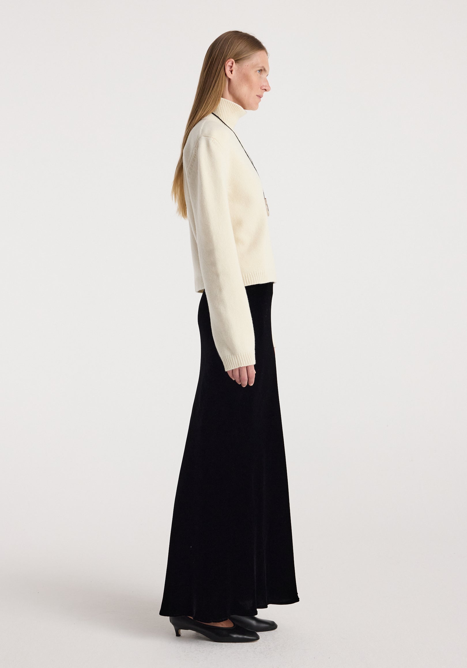 Woman in profile wearing a cream turtleneck sweater paired with a long black Fluid velvet skirt | black and black low-heeled shoes against a plain white background.