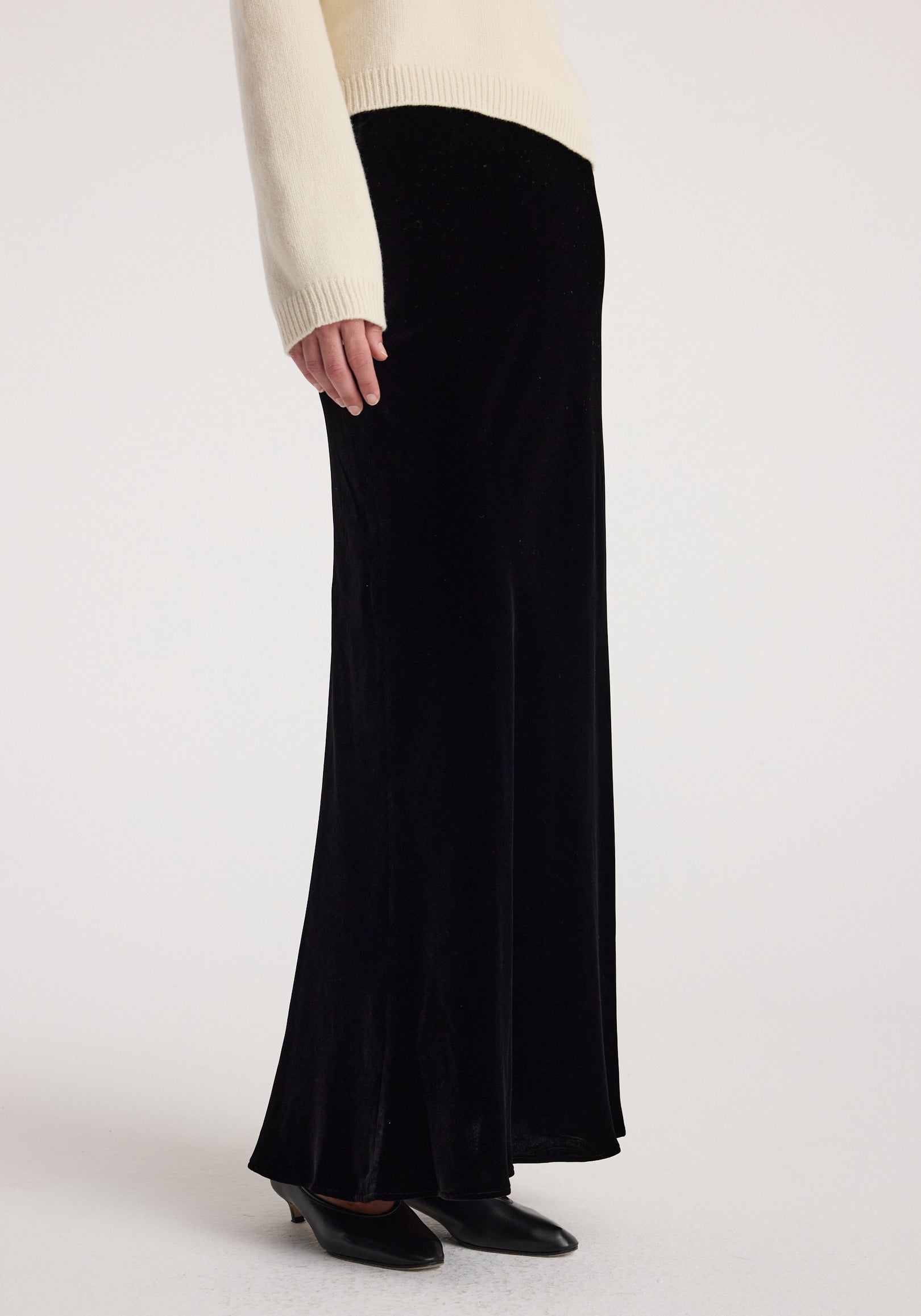 Side view of a person wearing a long black skirt made of smooth velvet fabric paired with black pointed shoes and a cream sweater, showcasing the Fluid velvet skirt | black.