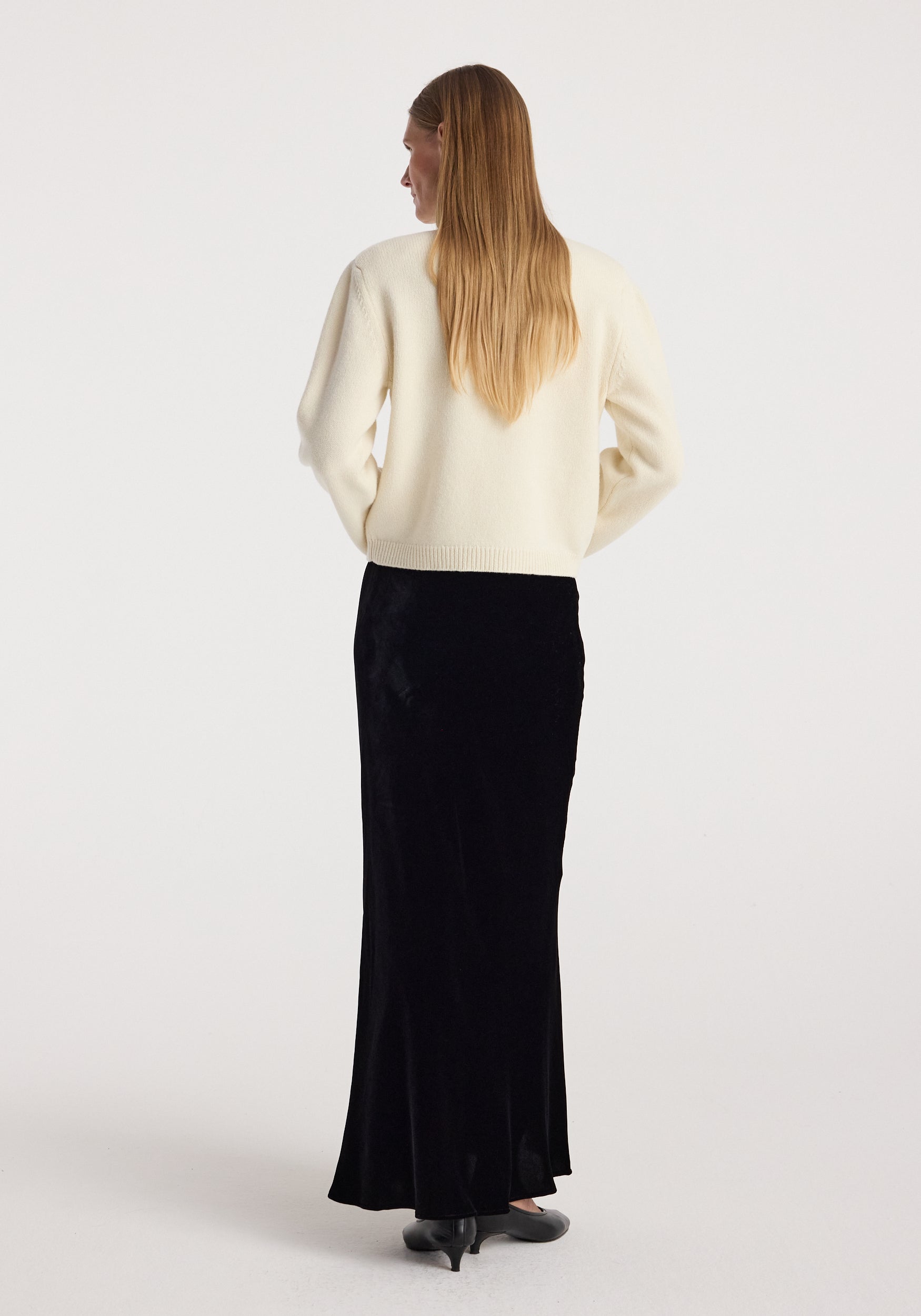 Woman wearing a black Fluid velvet skirt | black paired with a cream sweater, standing with her back to the camera against a plain light background.