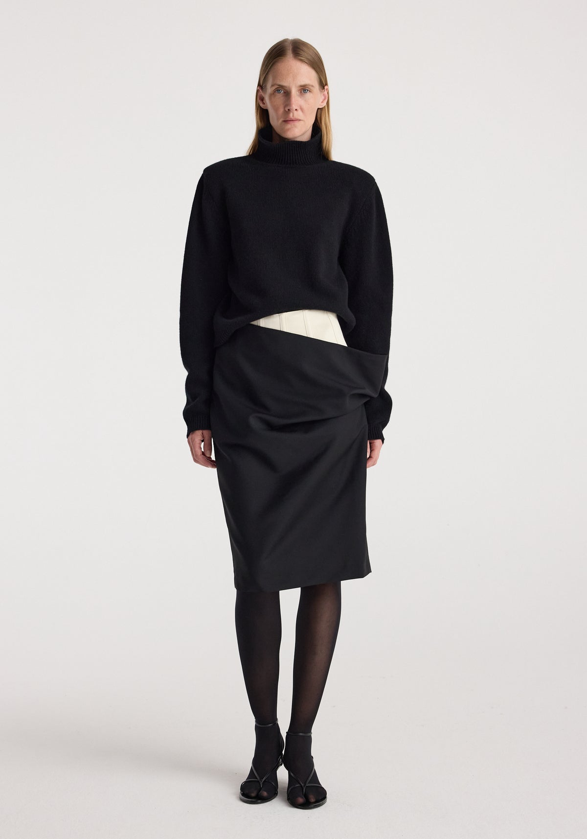 Model wearing a Double waistband skirt | black paired with a black long-sleeve turtleneck sweater, black sheer tights, and black open-toe heels against a plain white background.