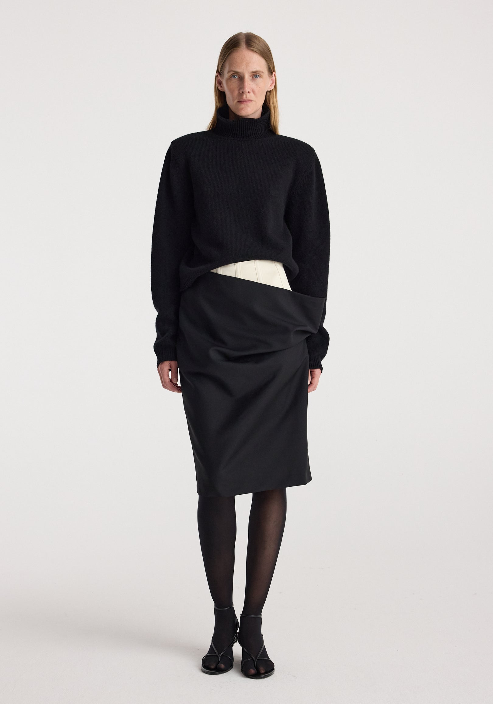 Model wearing a Double waistband skirt | black paired with a black long-sleeve turtleneck sweater, black sheer tights, and black open-toe heels against a plain white background.