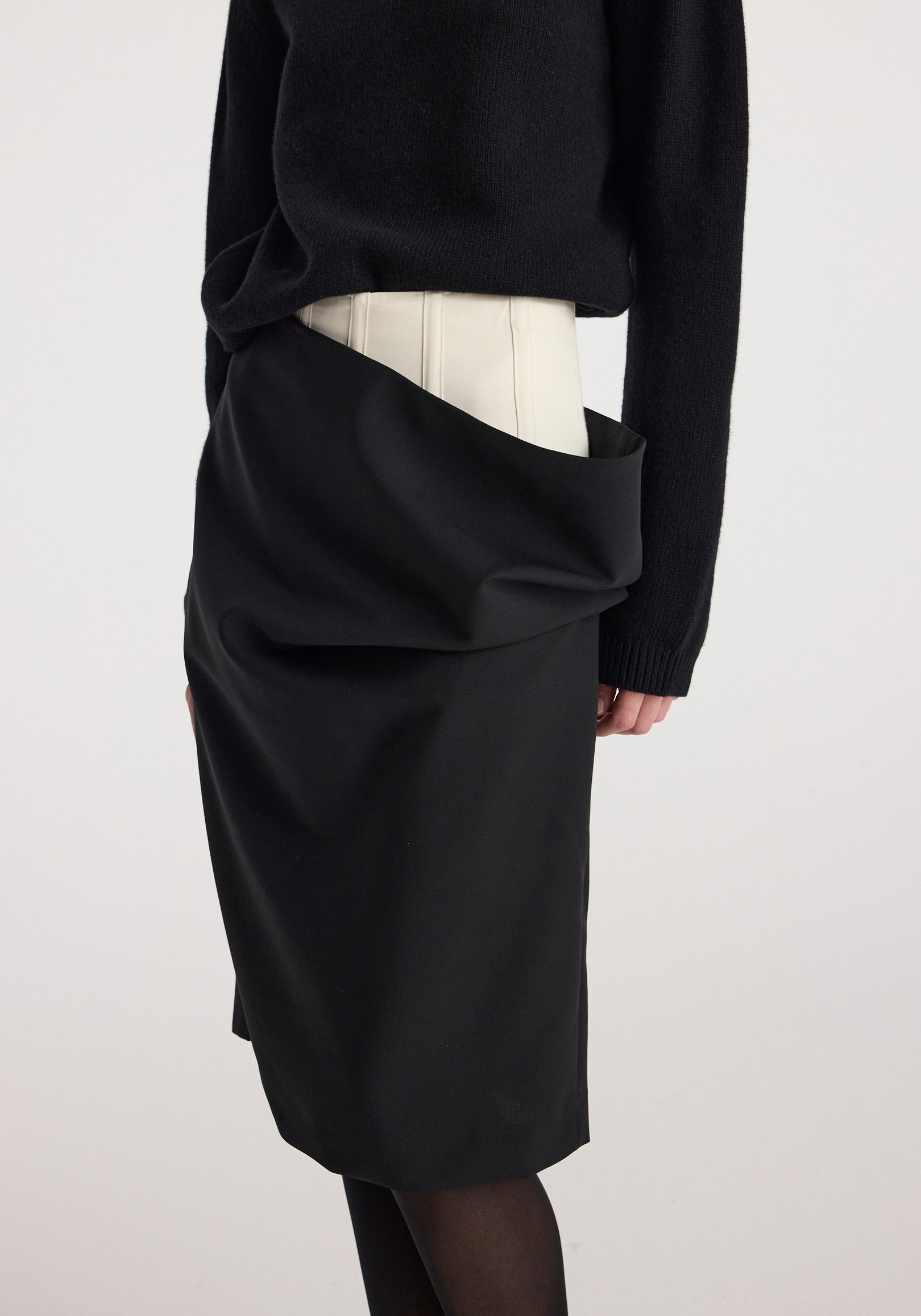 Close-up of a model wearing a Double waistband skirt | black, featuring a unique double waistband design with an outer black layer draped over a lighter inner waistband, paired with a black sweater.