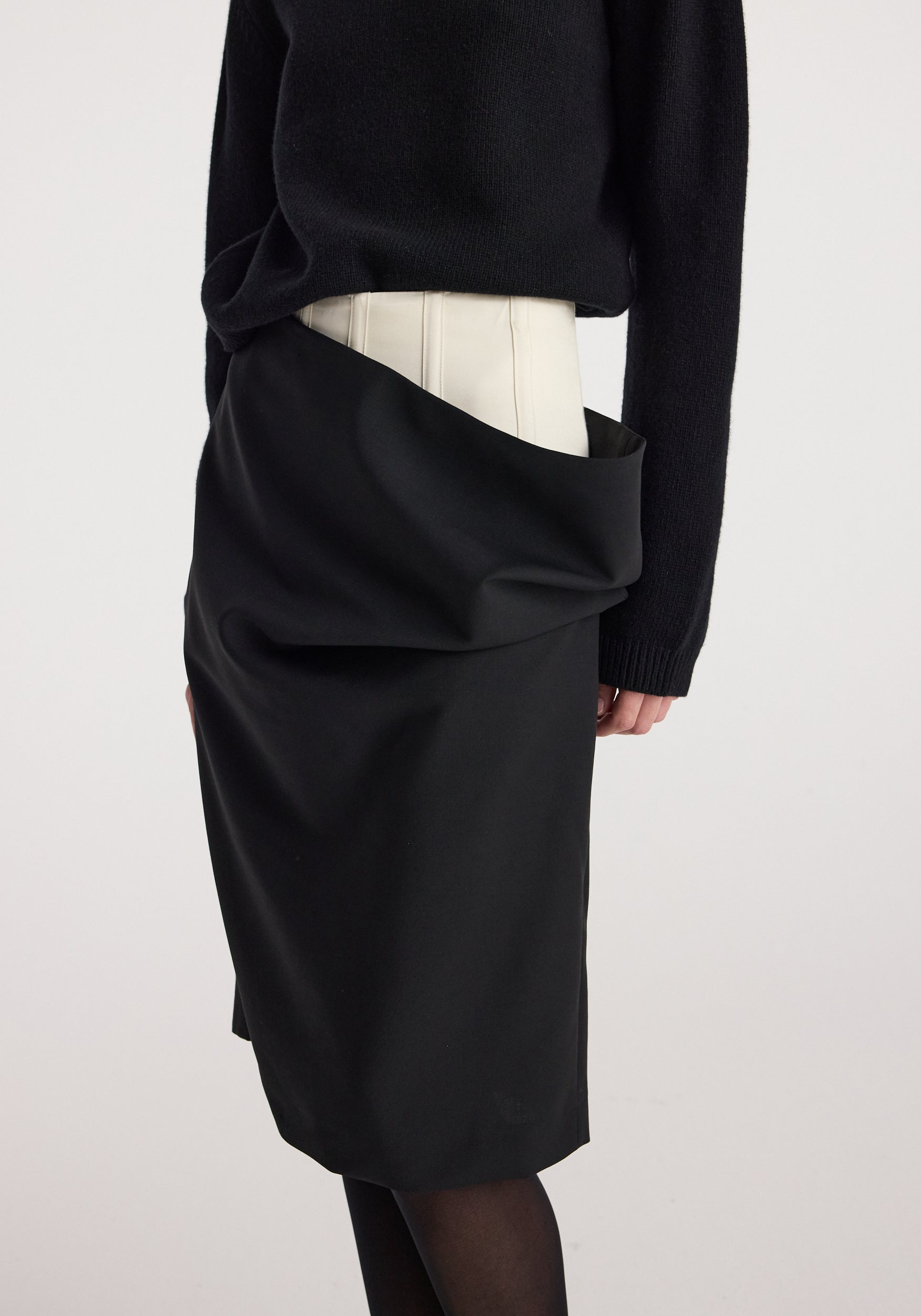 Close-up of a model wearing a Double waistband skirt | black, featuring a unique double waistband design with an outer black layer draped over a lighter inner waistband, paired with a black sweater.