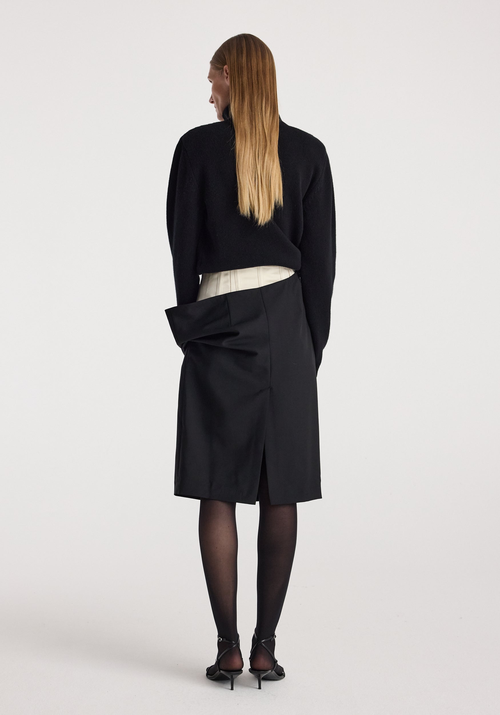 Woman wearing a black long-sleeve top and a Double waistband skirt | black with a white inner waistband detail, black tights, and black heels shown from the back.