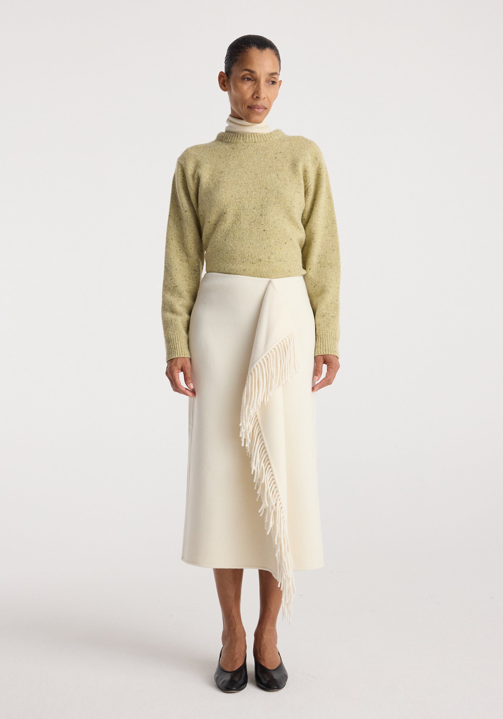 Woman wearing a light green sweater and a Double-faced fringe skirt | off white with asymmetrical fringe detail, paired with black shoes against a plain background.