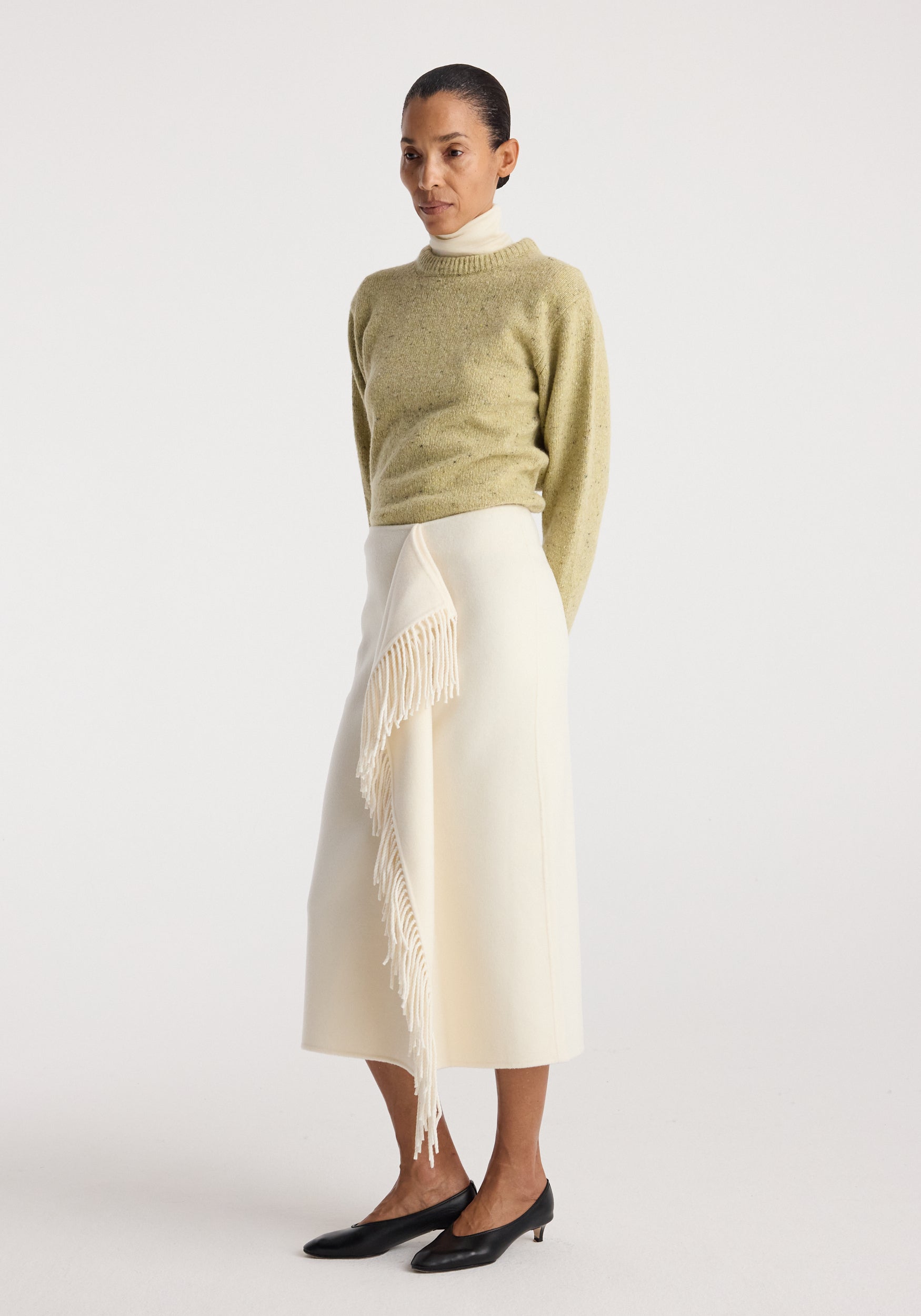 Woman wearing a light green sweater and a Double-faced fringe skirt | off white with layered fringe detail, standing against a plain background with black low-heeled shoes.