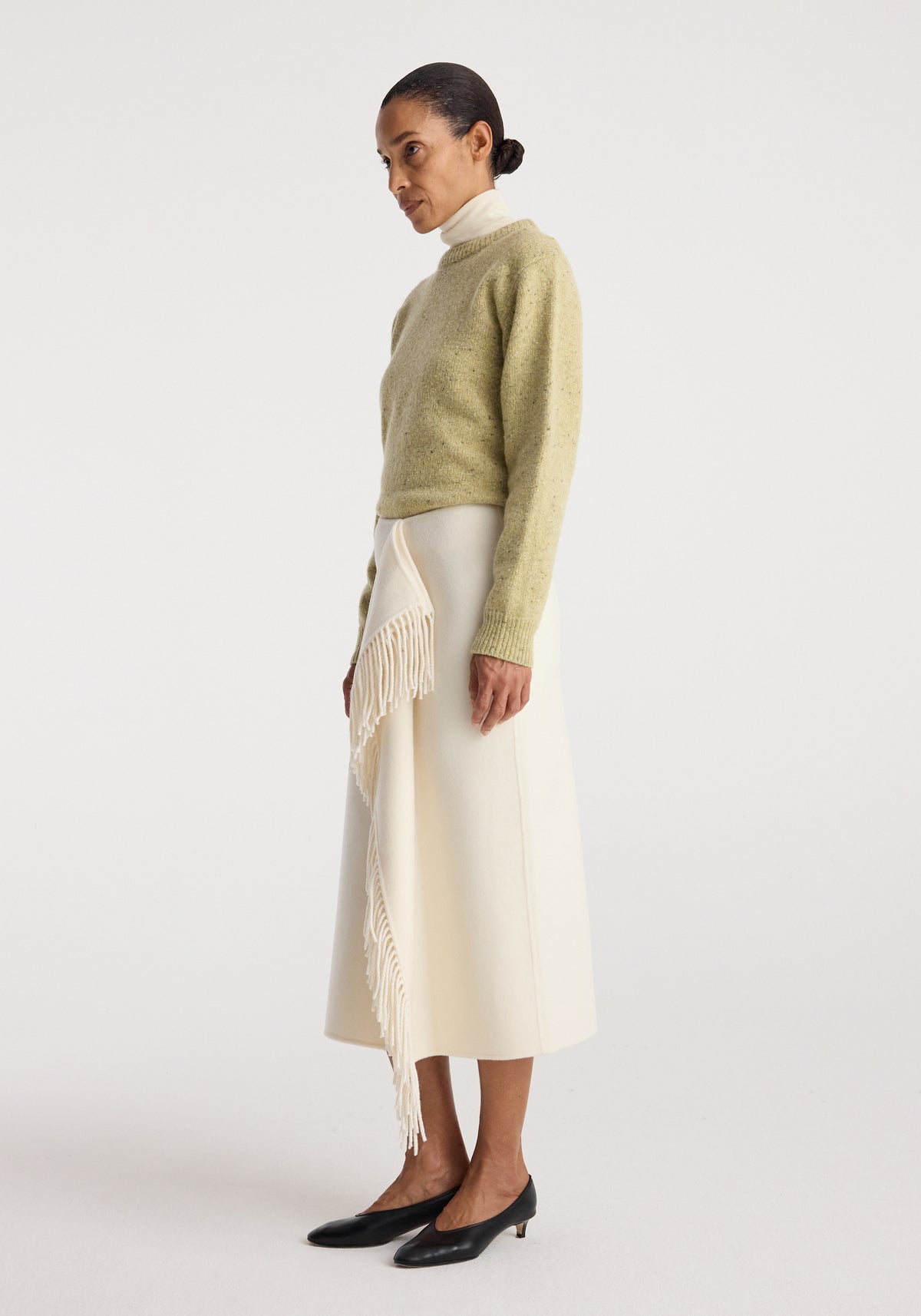 Woman wearing a light green sweater paired with a Double-faced fringe skirt | off white, featuring cascading fringe detail on one side, styled with black low heels.