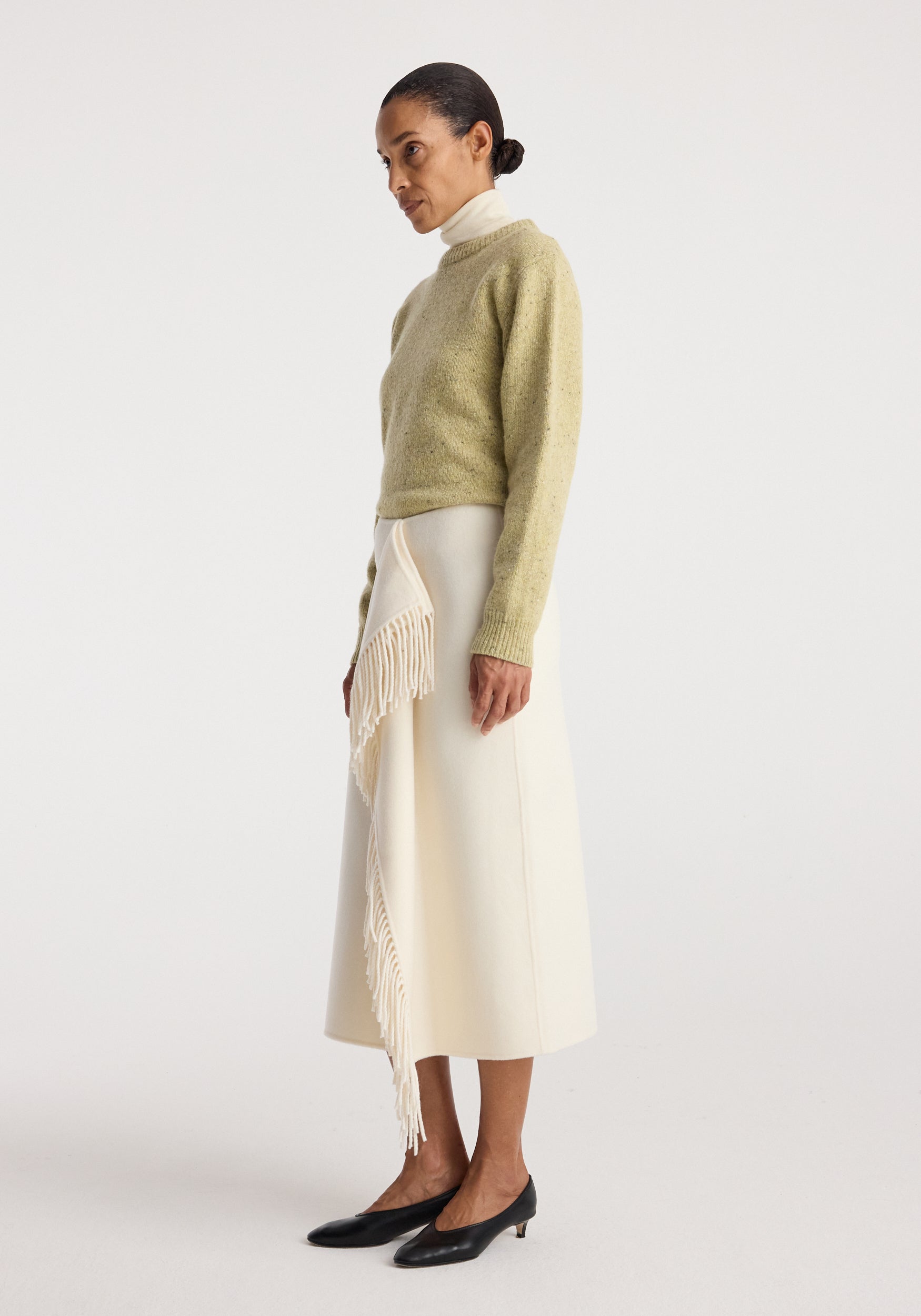 Woman wearing a light green sweater paired with a Double-faced fringe skirt | off white, featuring cascading fringe detail on one side, styled with black low heels.