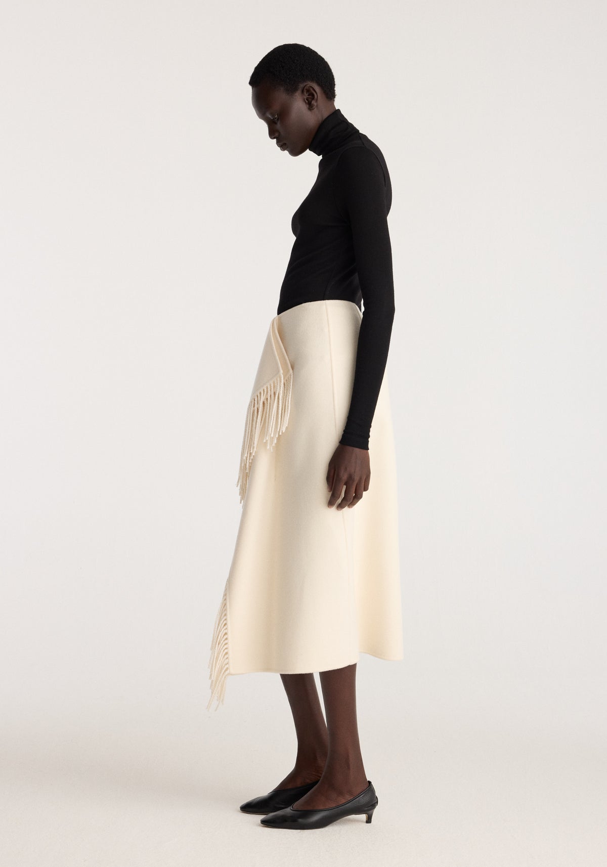 Double-faced fringe skirt | off white styled with a black fitted top and pointed heels, featuring a midi length and cascading fringe details along the sides and hem