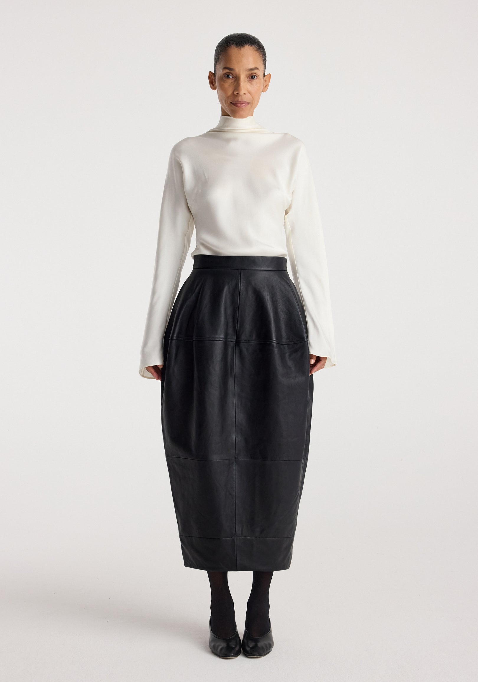 Woman wearing a fitted high-waisted Sculptural leather skirt | black paired with a long-sleeve white top and black shoes, standing against a plain background.