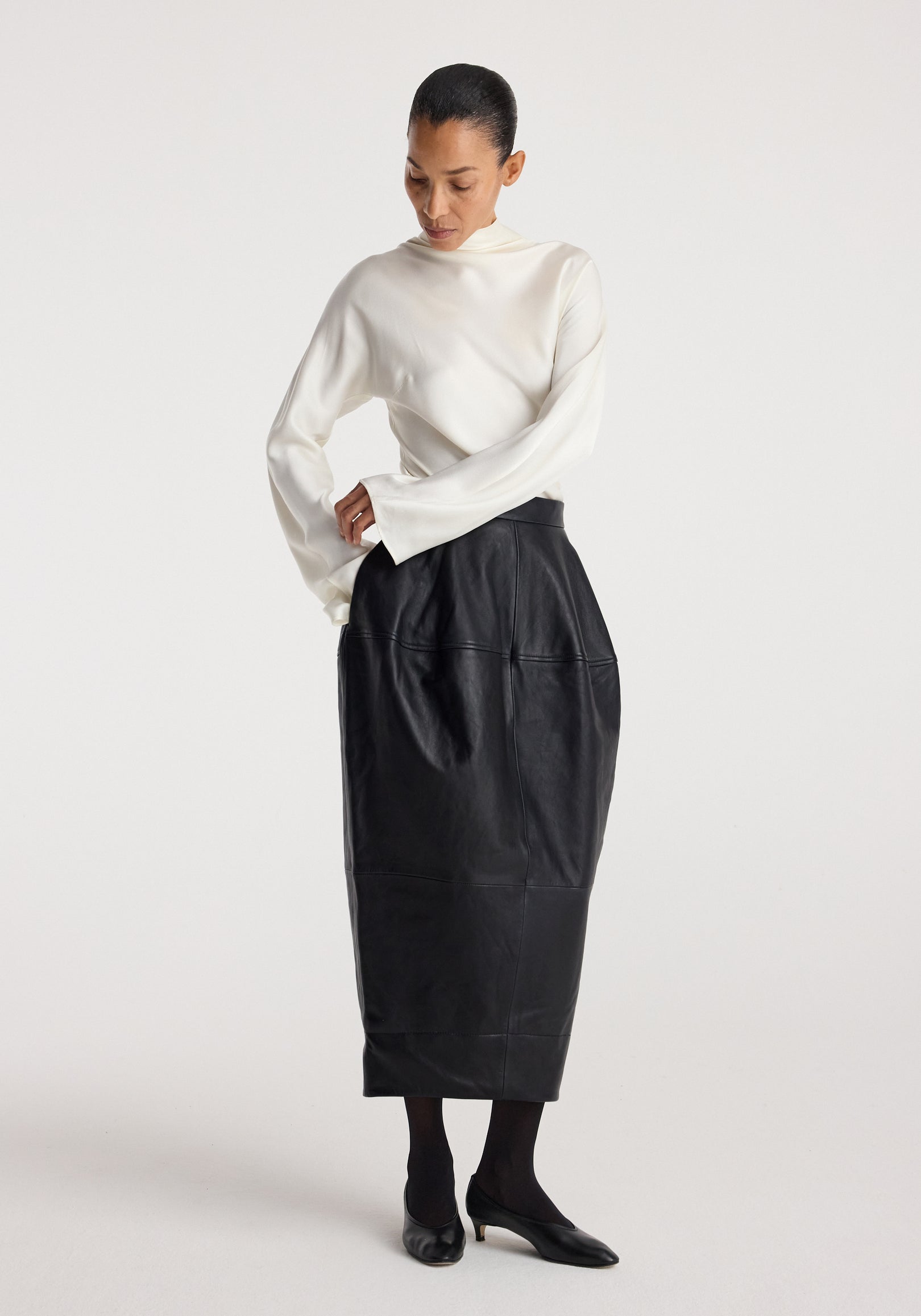 Woman wearing a Sculptural leather skirt | black with a high waist and geometric panel details, paired with a white long-sleeve blouse and black heels on a light background.