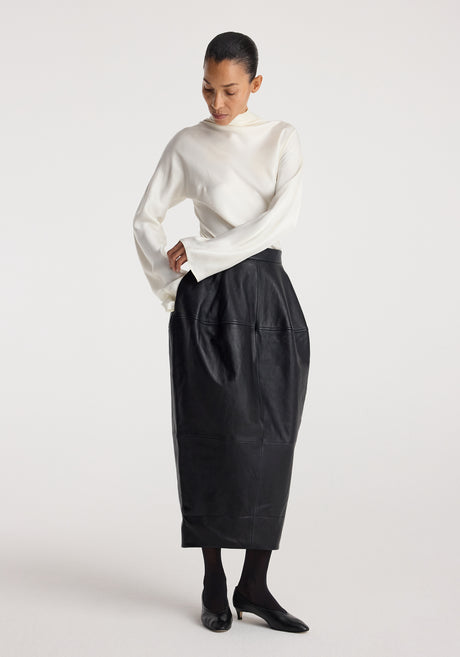 Woman wearing a fitted high-waisted Sculptural leather skirt | black paired with a long-sleeve white top and black shoes, standing against a plain background.