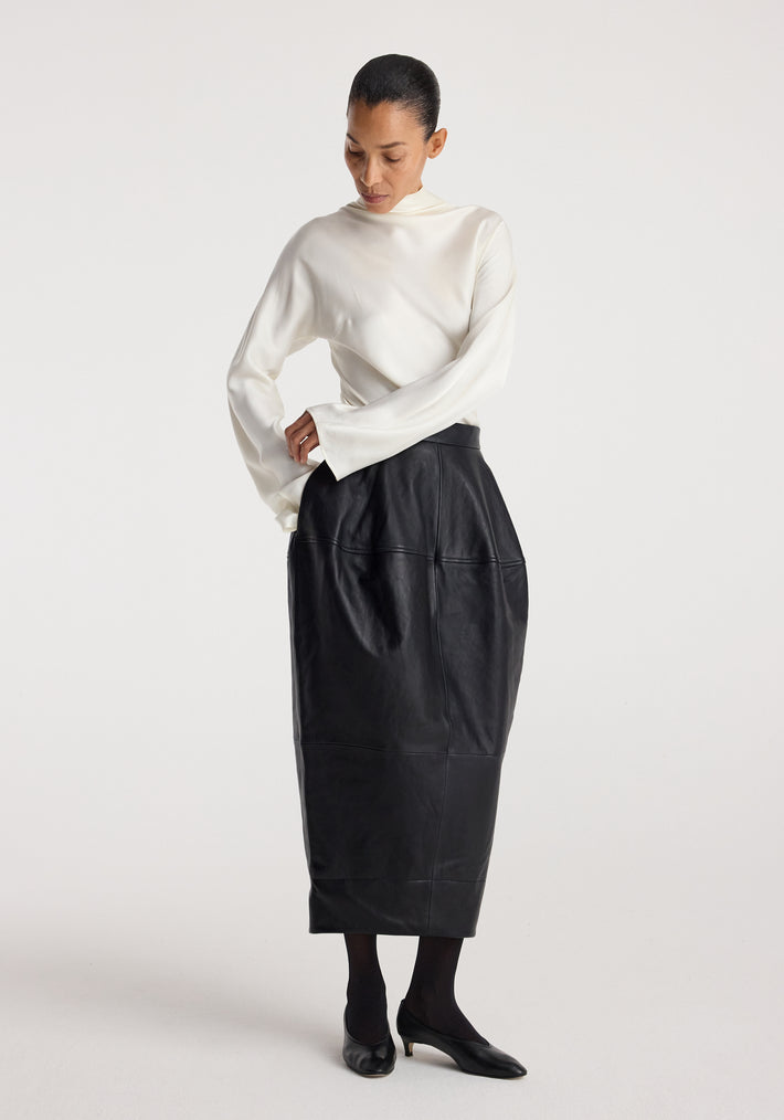Woman wearing a Sculptural leather skirt | black with a high waist and geometric panel details, paired with a white long-sleeve blouse and black heels on a light background.