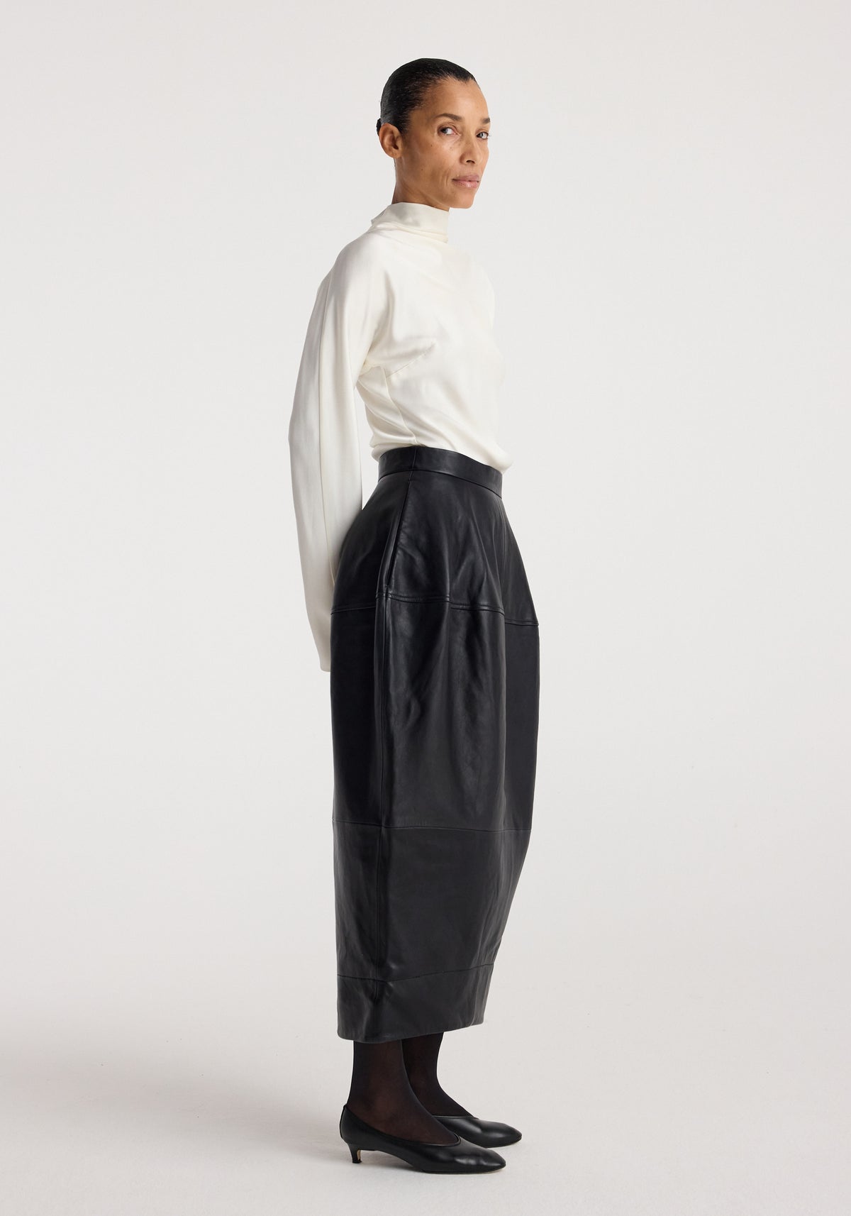 Woman wearing a white long-sleeve top paired with a Sculptural leather skirt | black that has a high waist and a midi length, styled with black sheer tights and low black shoes.
