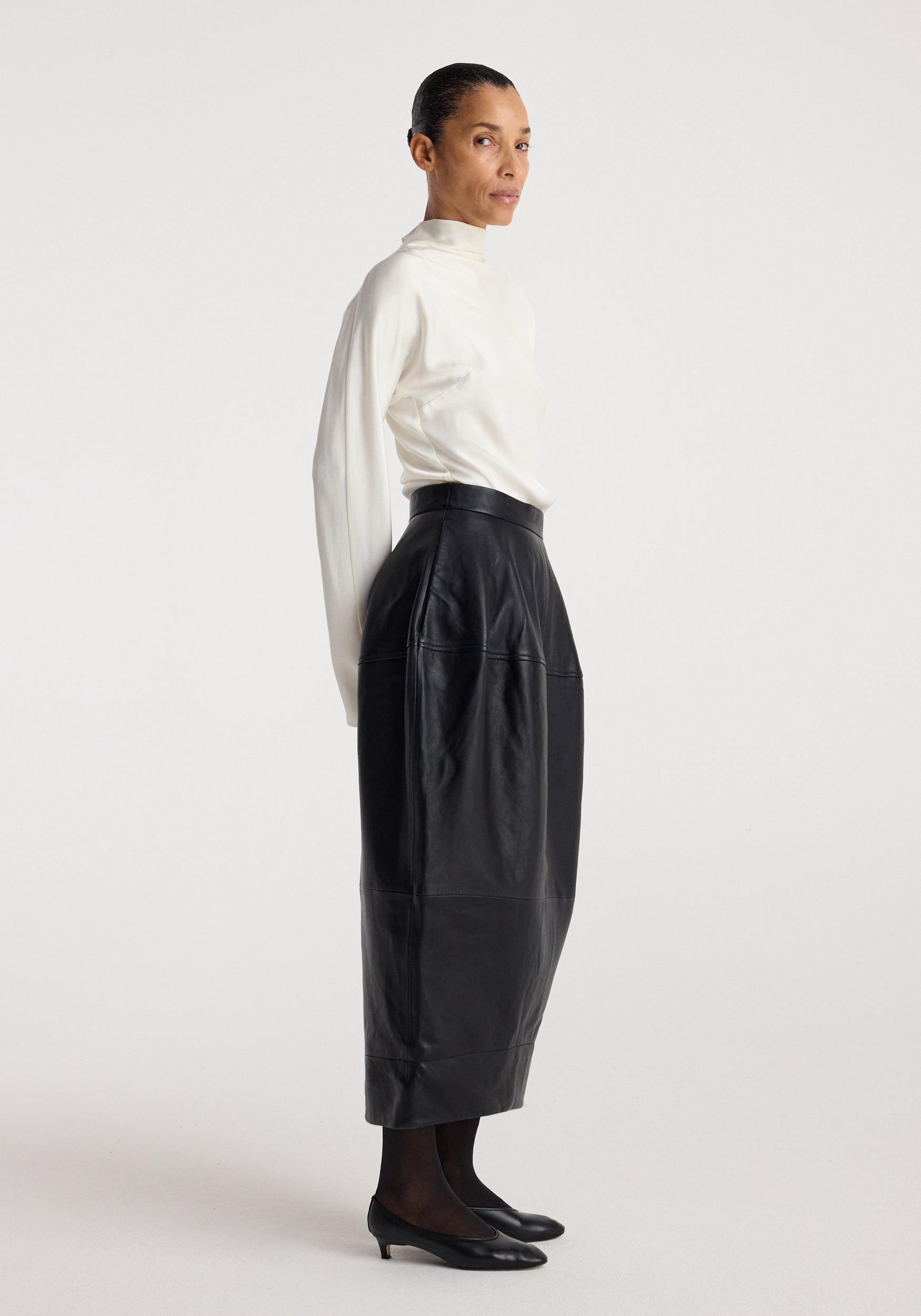 Woman wearing a white long-sleeve top paired with a Sculptural leather skirt | black that has a high waist and a midi length, styled with black sheer tights and low black shoes.