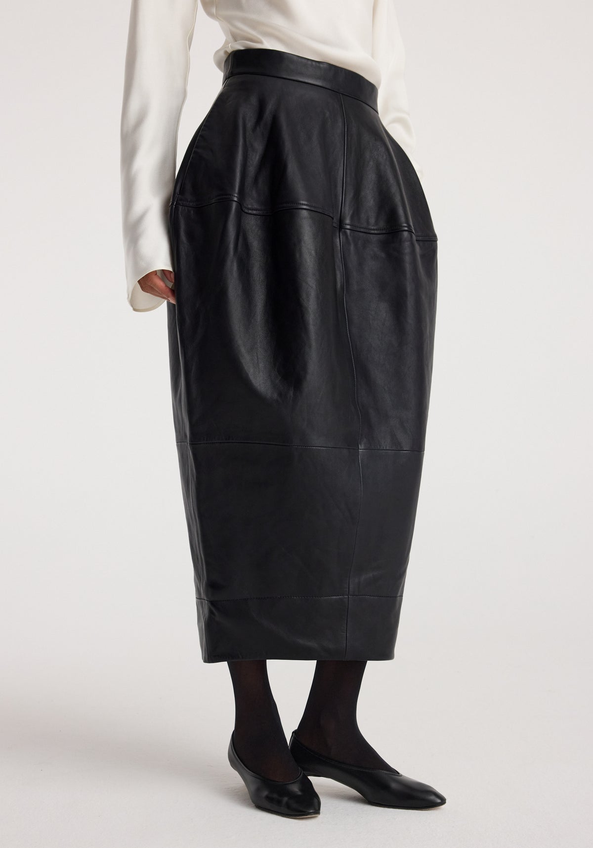 Woman wearing a black sculptural leather skirt | black with a high waist, paired with black tights and black flats against a plain background.