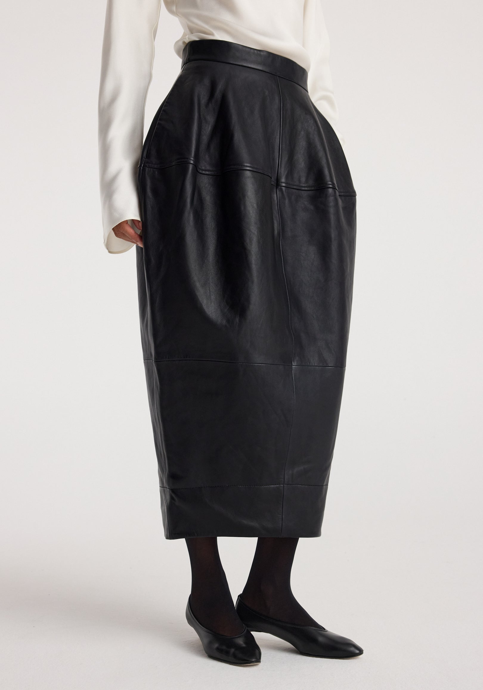 Woman wearing a black sculptural leather skirt | black with a high waist, paired with black tights and black flats against a plain background.