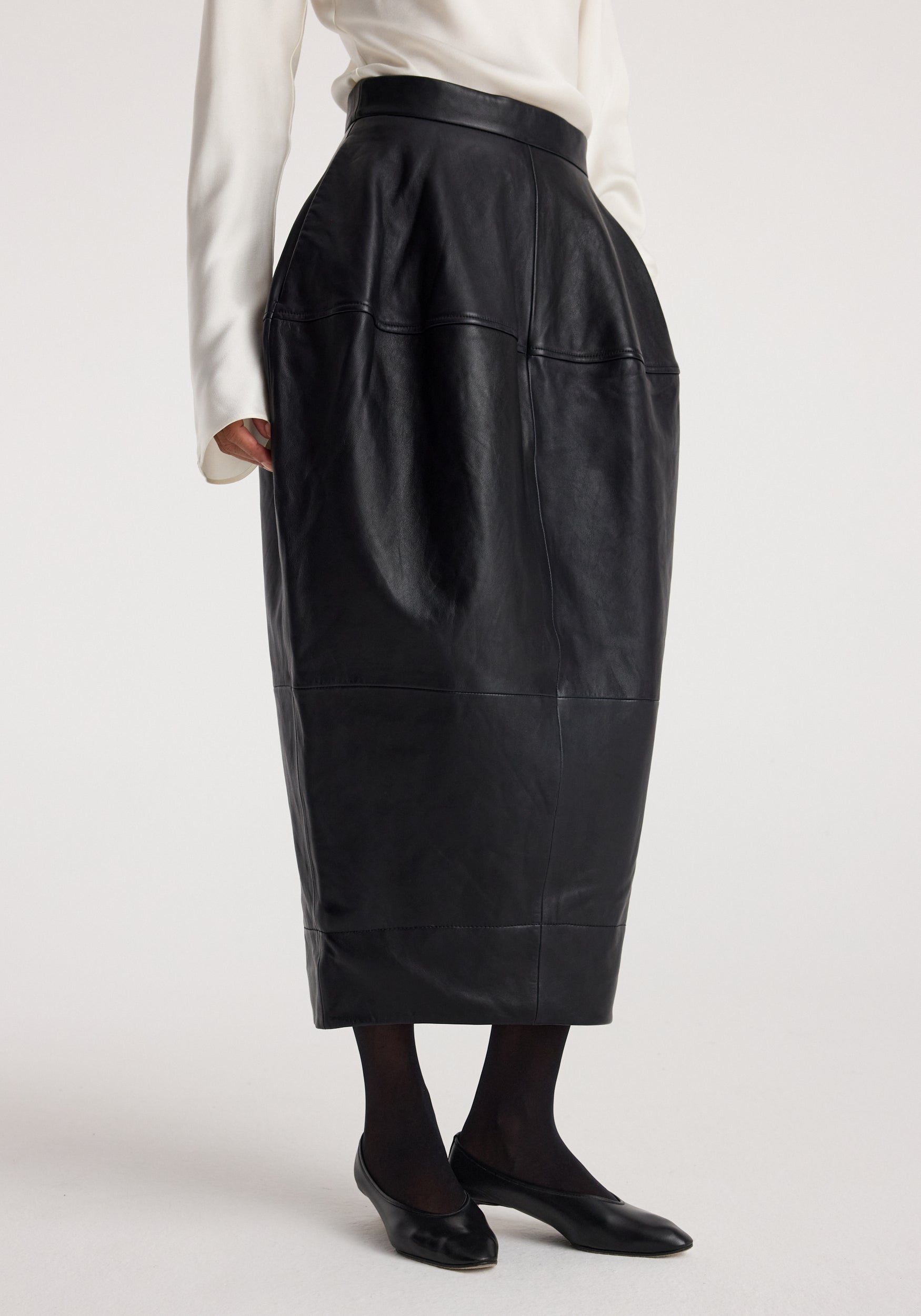 Woman wearing a black sculptural leather skirt | black with a high waist, paired with black tights and black flats against a plain background.