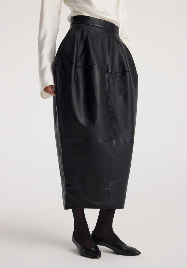 Woman wearing a black sculptural leather skirt | black with a high waist, paired with black tights and black flats against a plain background.