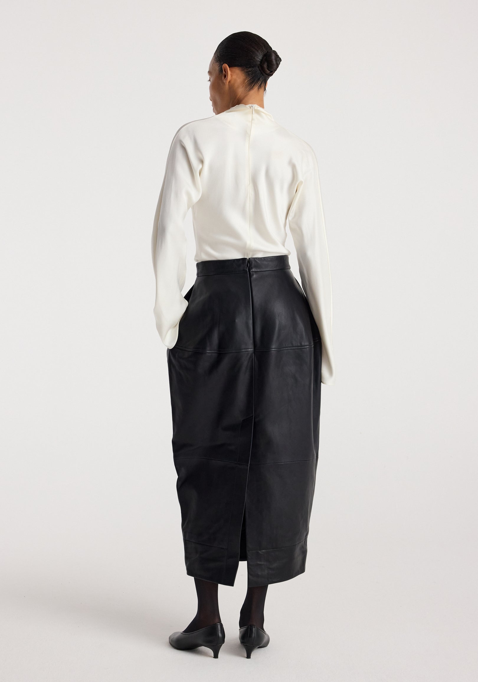 Woman wearing a long black Sculptural leather skirt | black paired with a white top and black heels, standing against a plain light background shown from the back.
