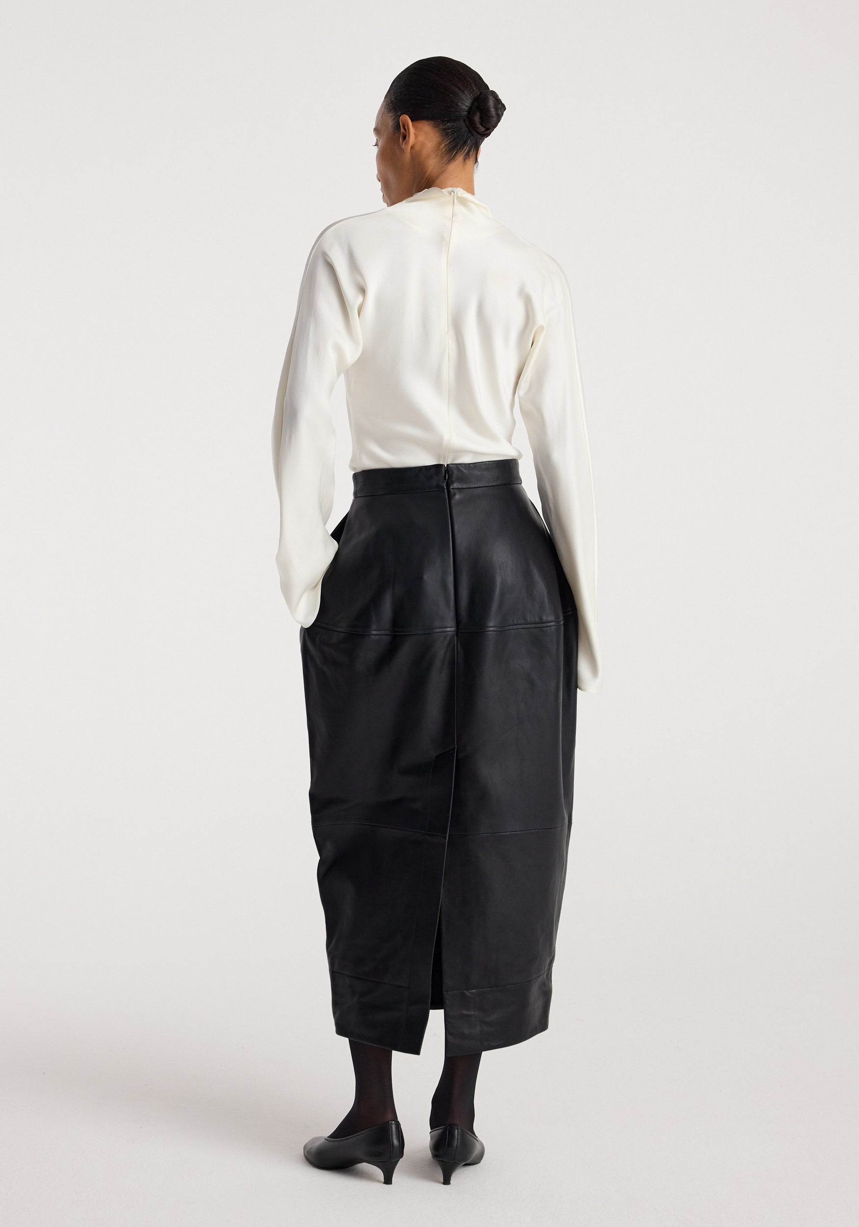 Woman wearing a long black Sculptural leather skirt | black paired with a white top and black heels, standing against a plain light background shown from the back.