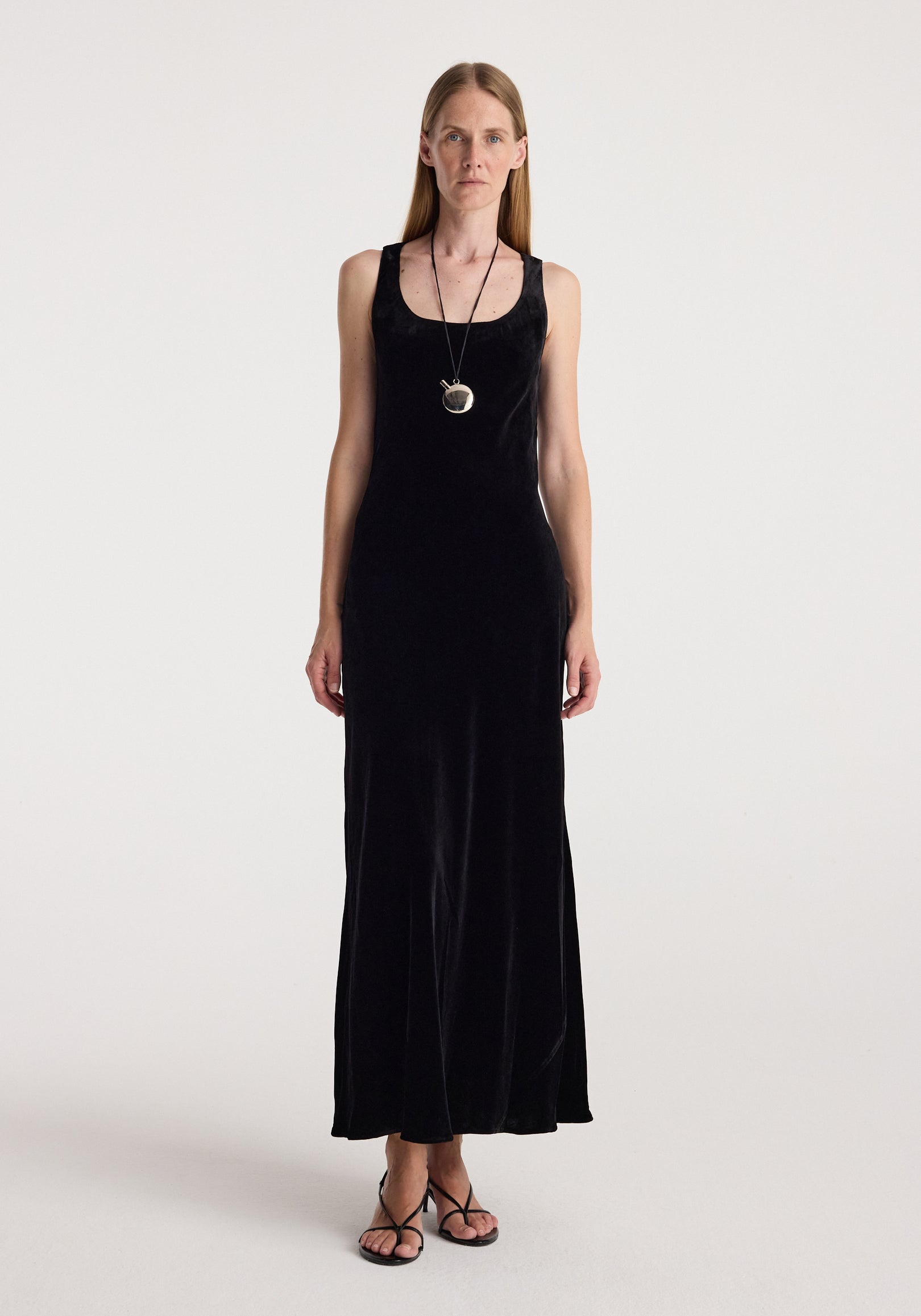 Woman wearing a long black scoop-neck velvet dress | black with thin straps, paired with a pendant necklace and black strappy sandals, standing against a plain background.