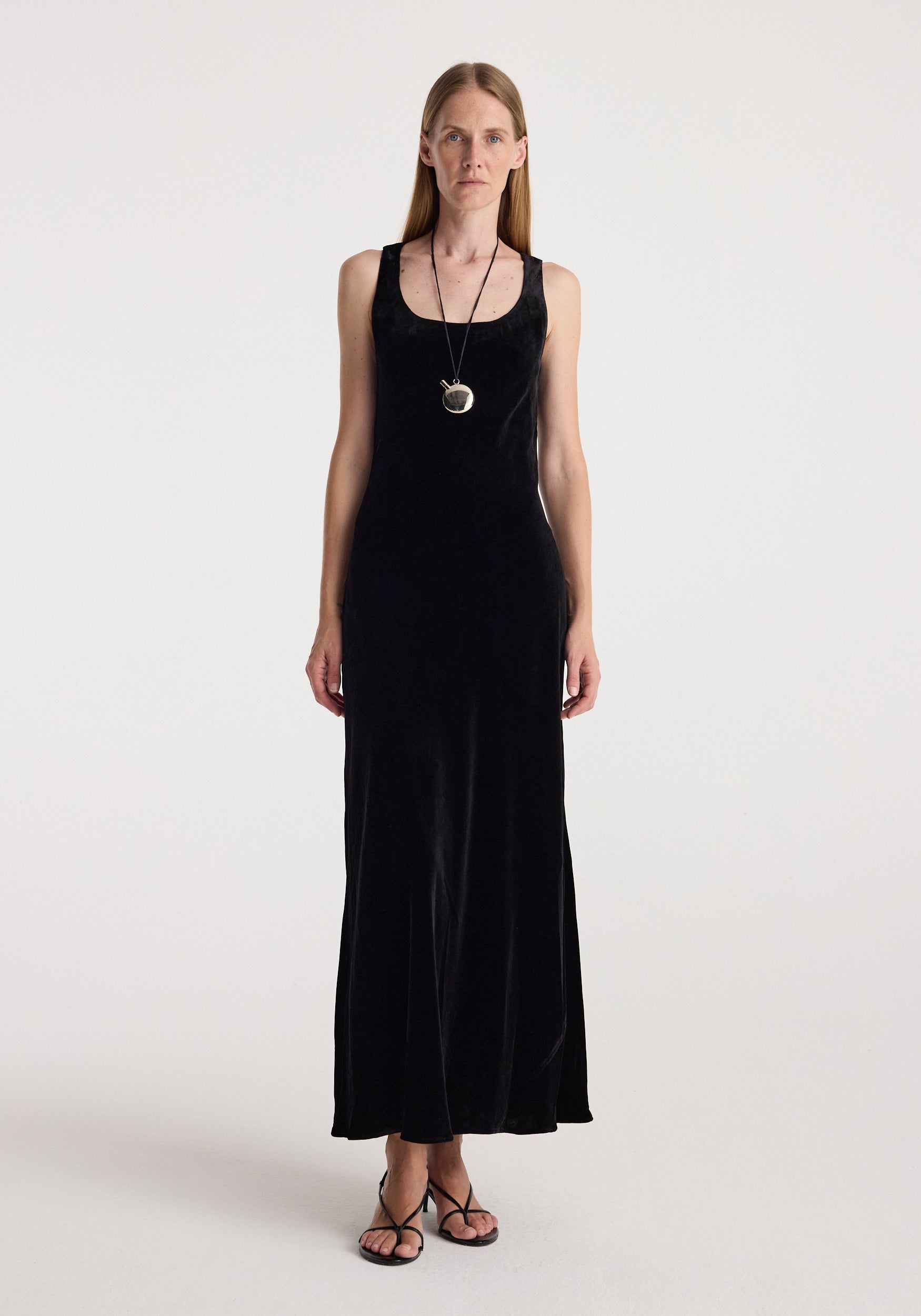 Woman wearing a long black scoop-neck velvet dress | black with thin straps, paired with a pendant necklace and black strappy sandals, standing against a plain background.