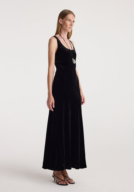 Woman wearing a long black scoop-neck velvet dress | black with thin straps, paired with a pendant necklace and black strappy sandals, standing against a plain background.